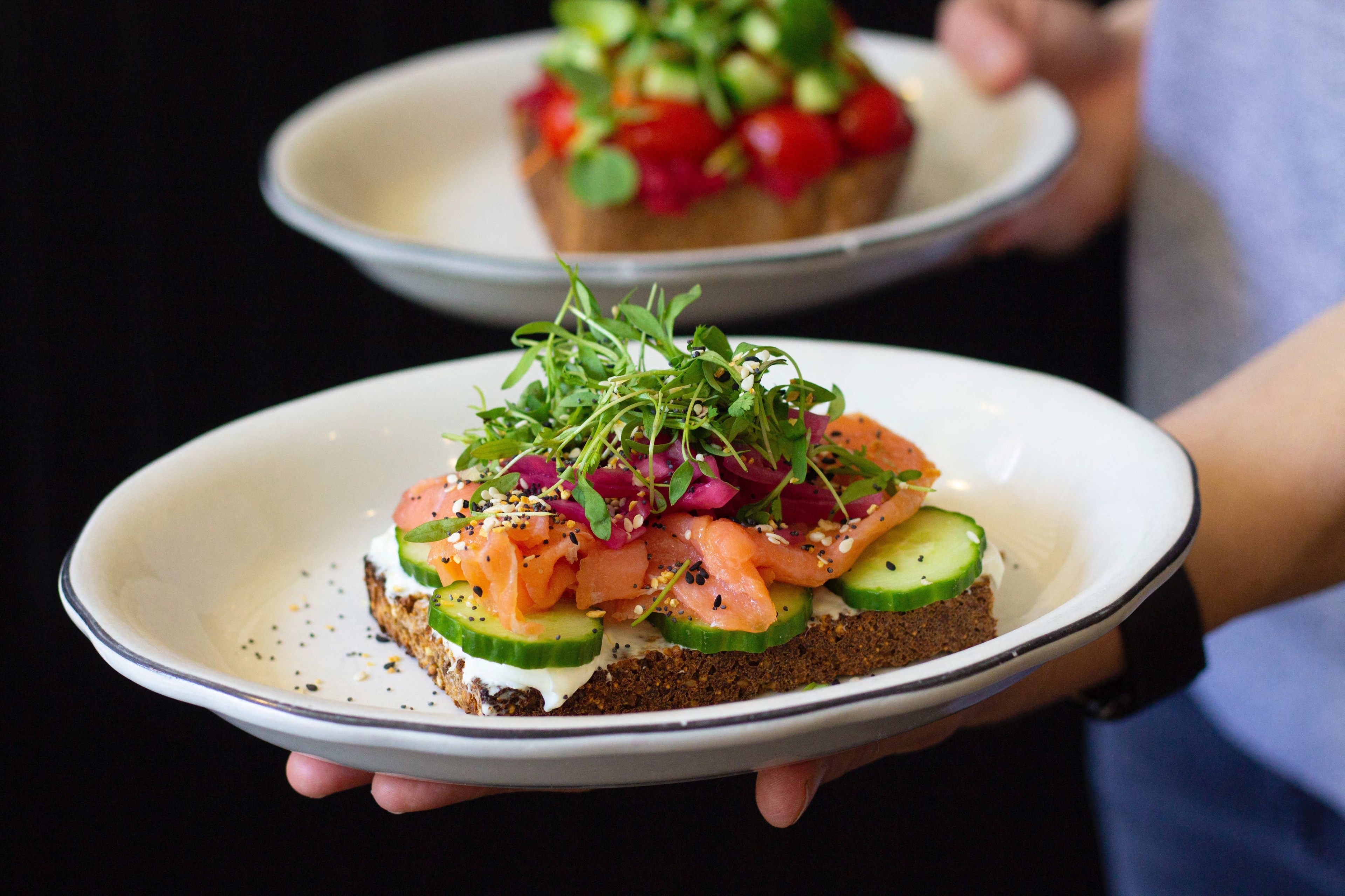 A person holds two plates of open-faced sandwiches. The front sandwich features smoked salmon, cucumber, greens, and seeds on dark bread; the back has colorful toppings.
