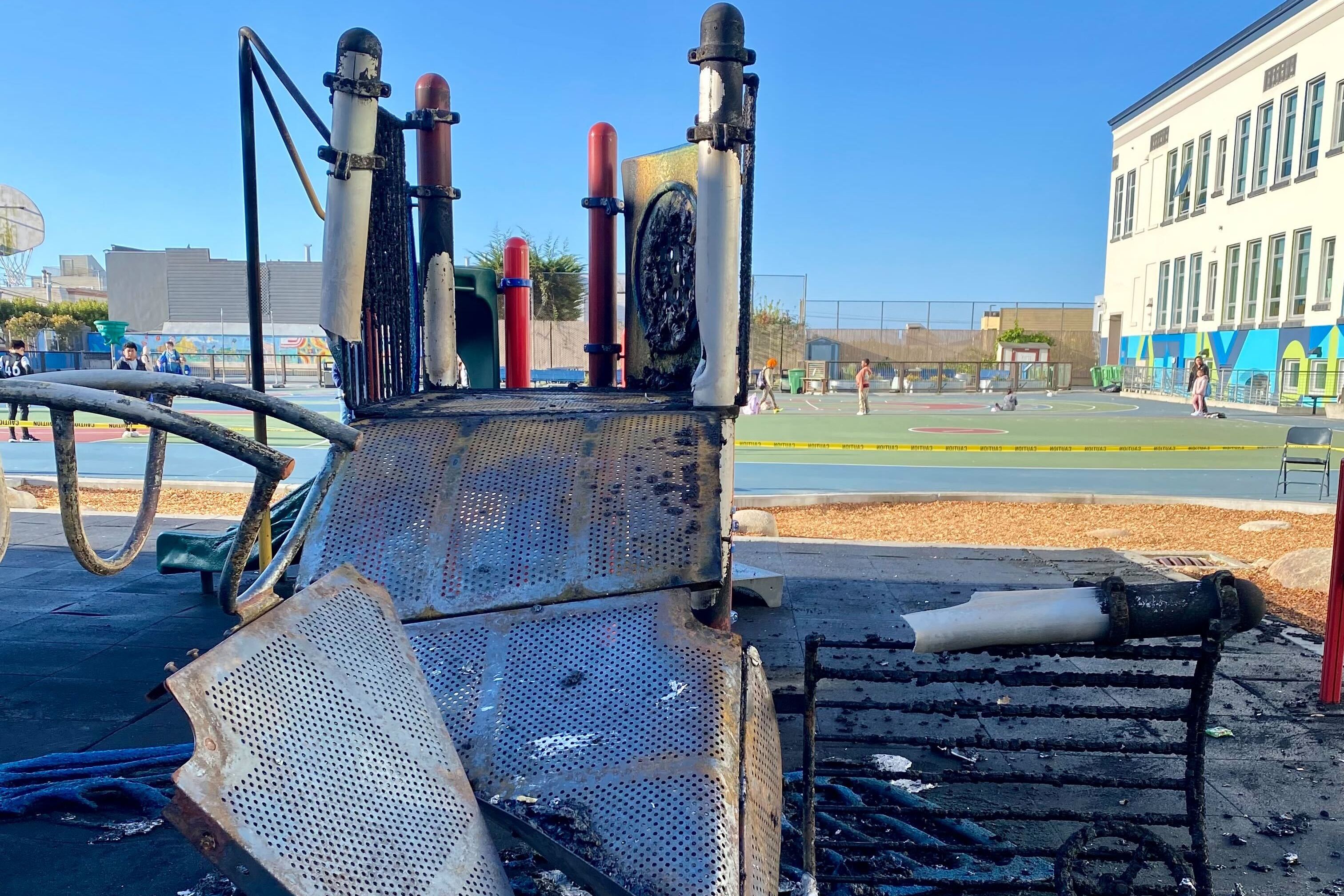 The image shows a burnt playground slide with melted plastic and metal debris. A school building and basketball court are visible in the background.