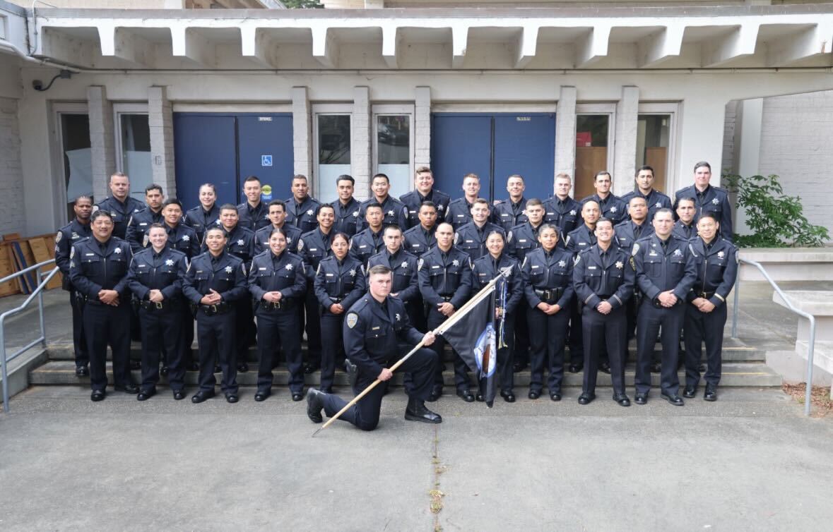A group of uniformed officers pose for a photo, with one kneeling in front holding a flag. They stand in front of a building with stairs on each side.