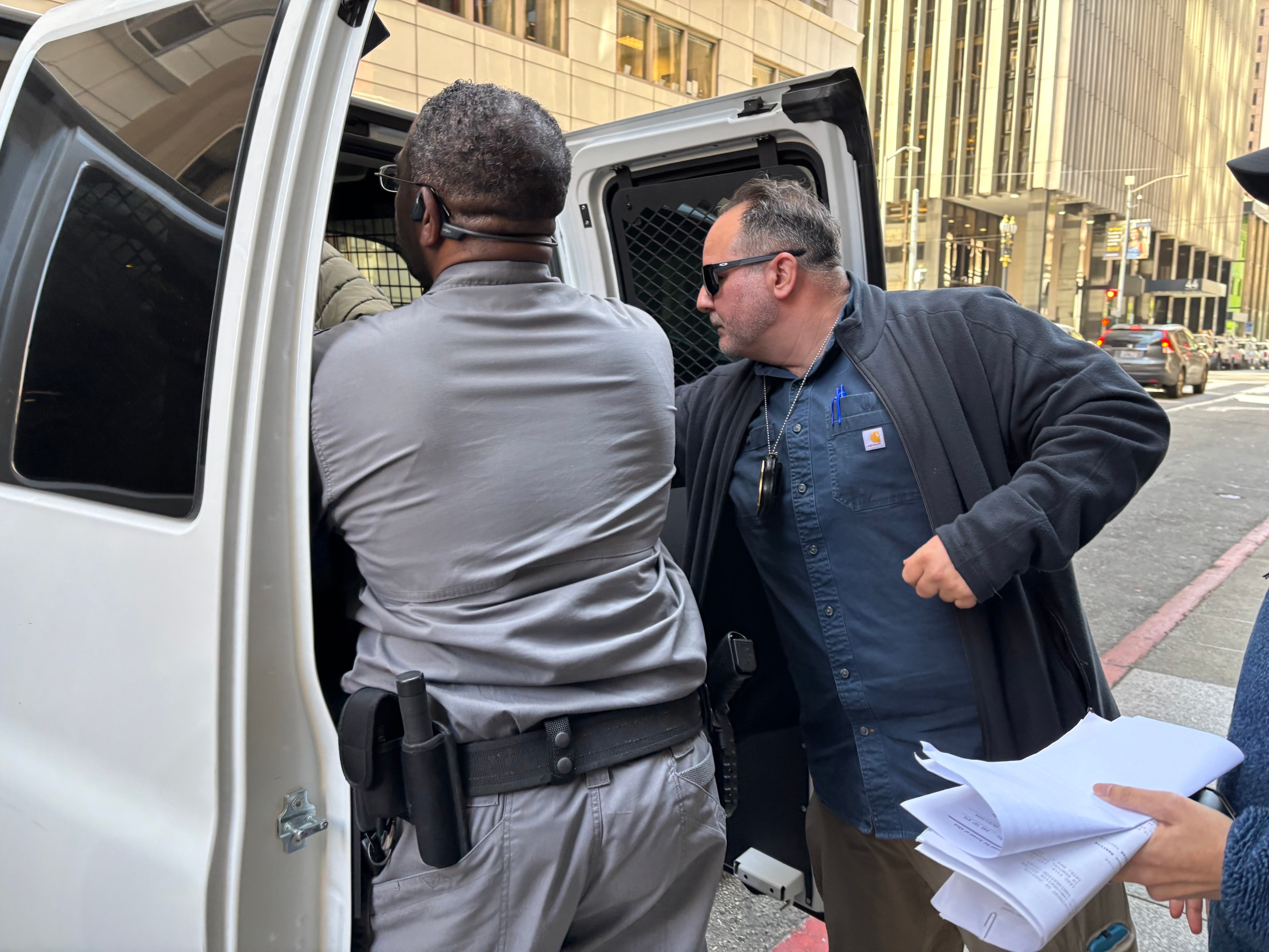 Two men are seen loading items into the back of a white van on a city street. One holds documents, and there's an office building in the background.