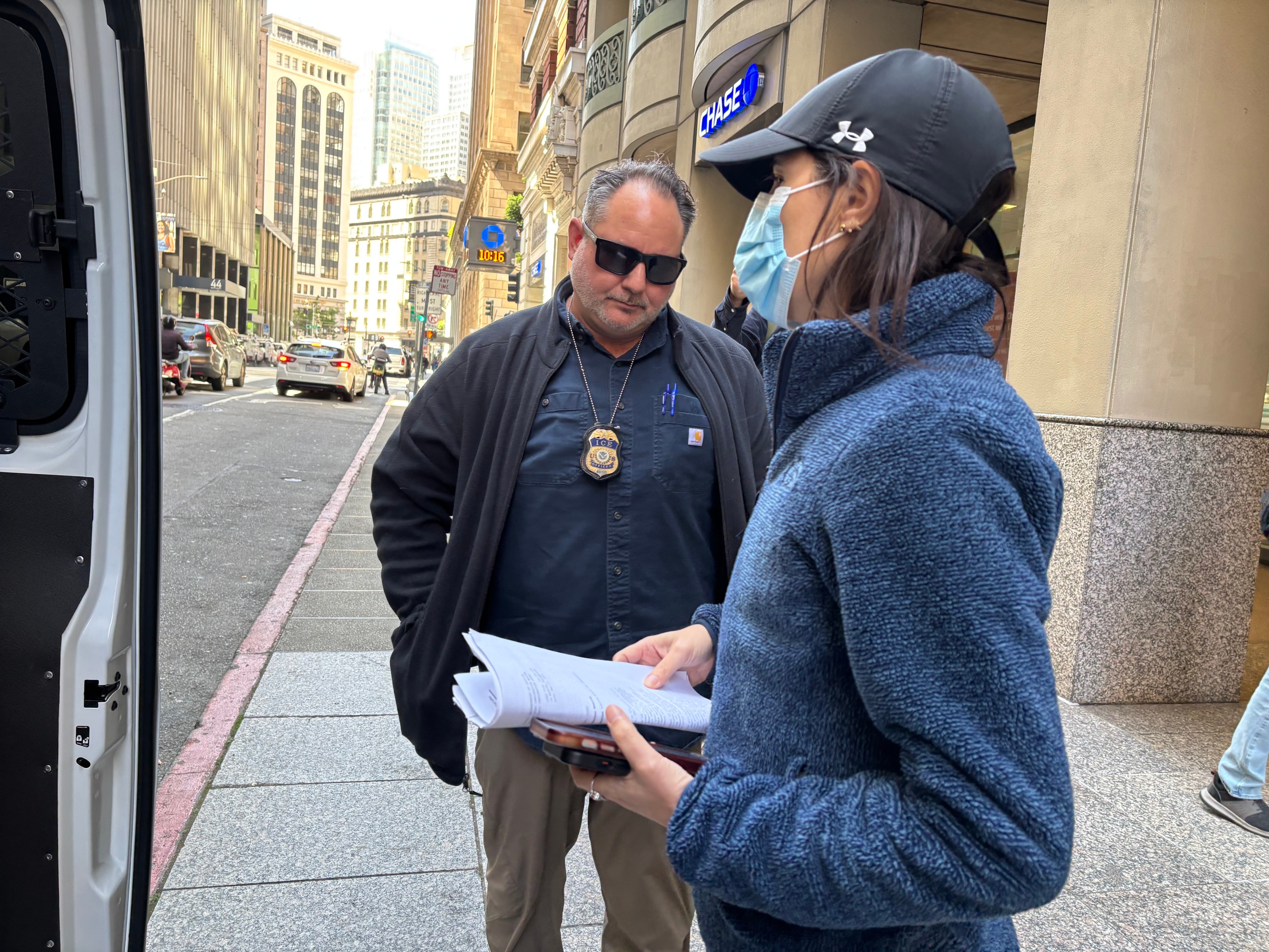 A man in dark clothing with a badge is talking to a woman in a blue jacket and cap, holding papers. They're on a city street near a Chase bank.