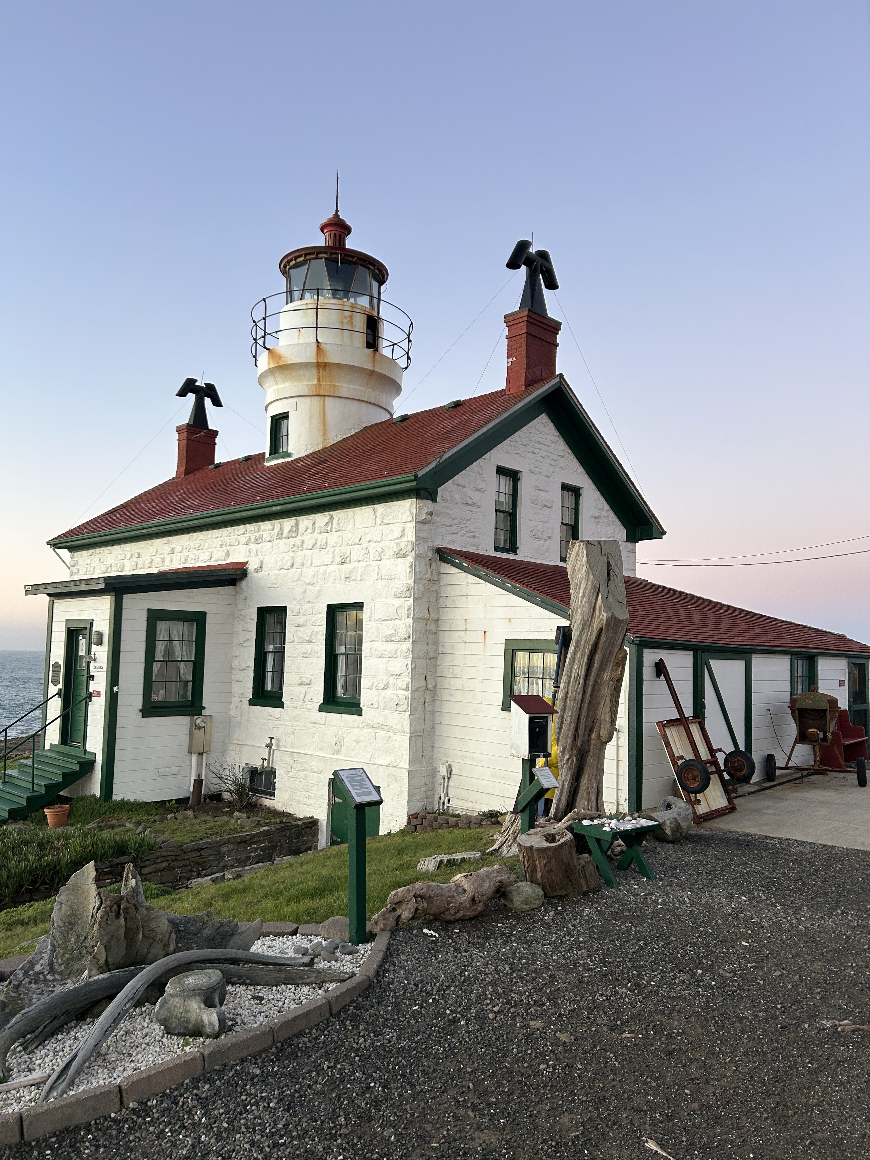 A white lighthouse with a red roof is situated by the ocean. The building features a tall lantern room and decorative rocks and wood pieces are placed in front.