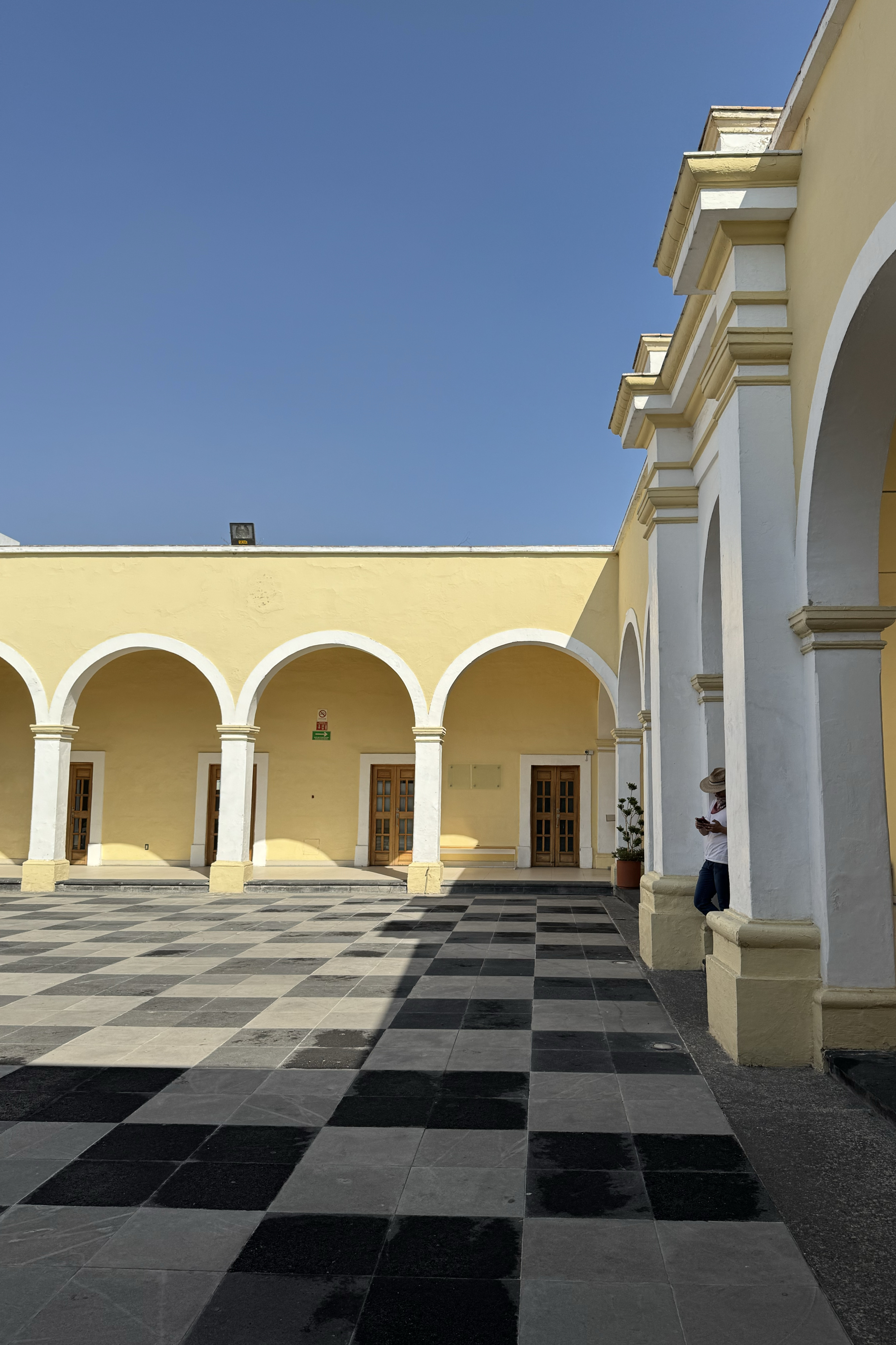 The image shows a sunny courtyard with yellow walls, white arches, and a checkered tile floor. A person leans against a column, looking at a phone.
