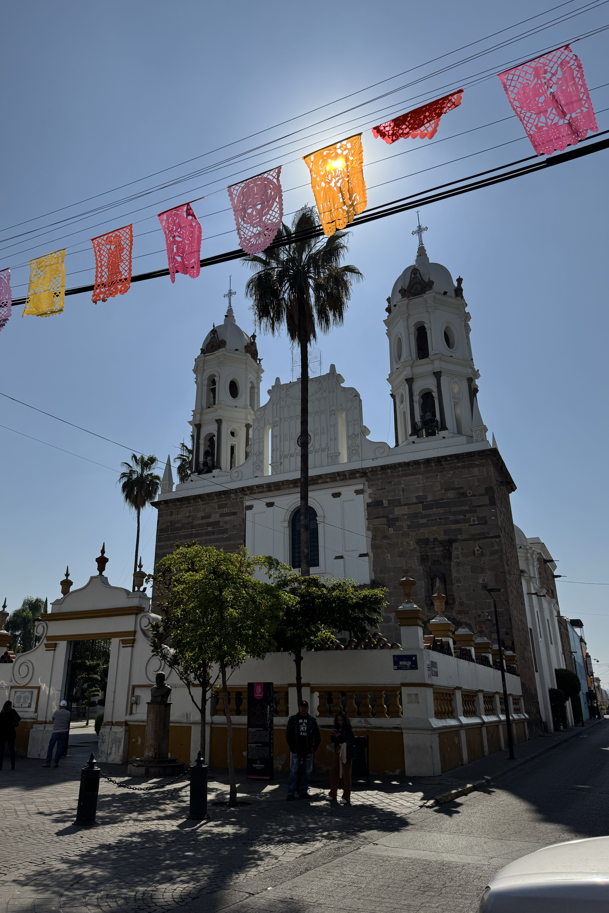 A large, historic church with twin towers stands under a clear blue sky. Colorful papel picado flags stretch above, and a palm tree is silhouetted against the sun.