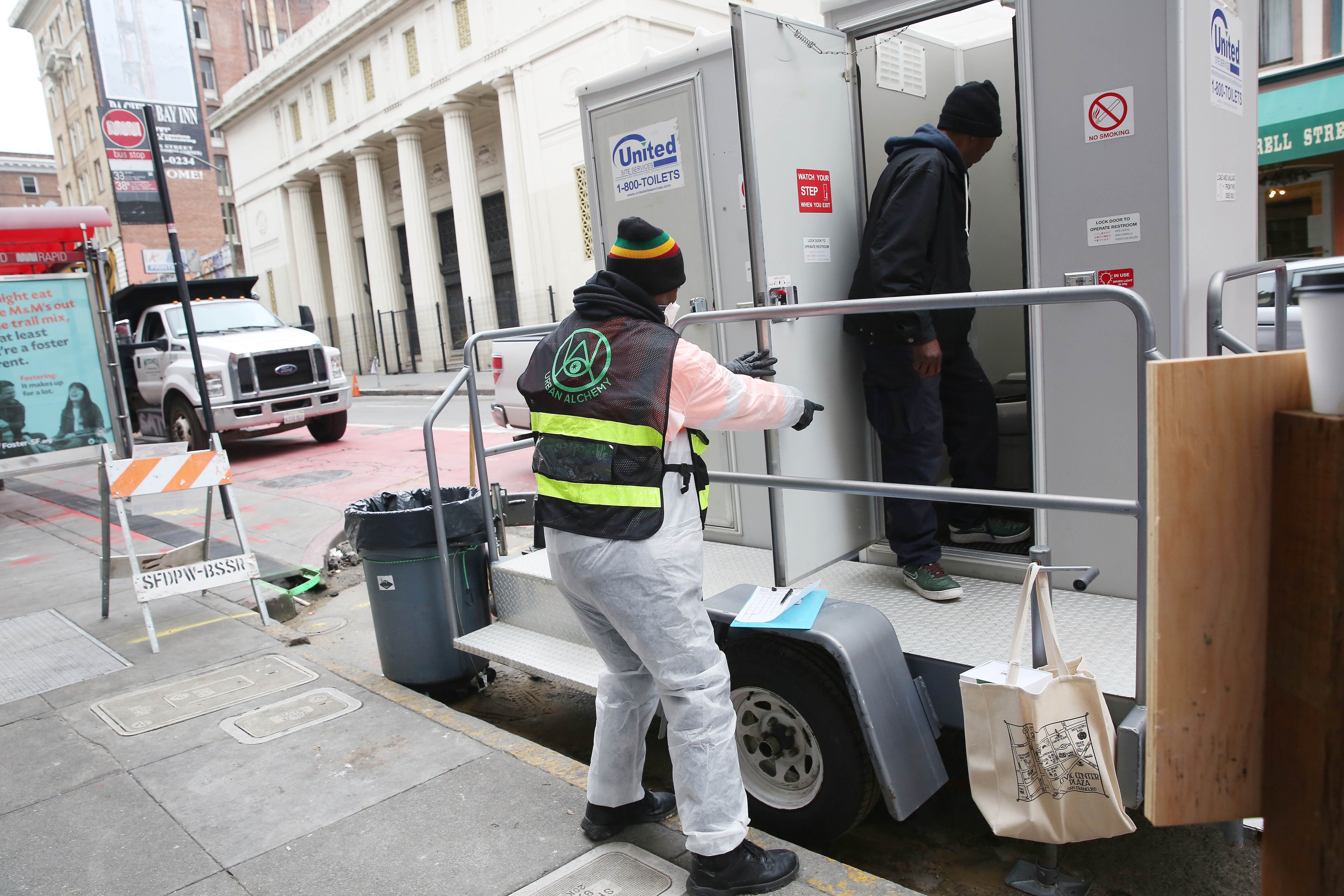 A uniformed Urban Alchemy practitioner stands near a portable toilet as someone enters it.