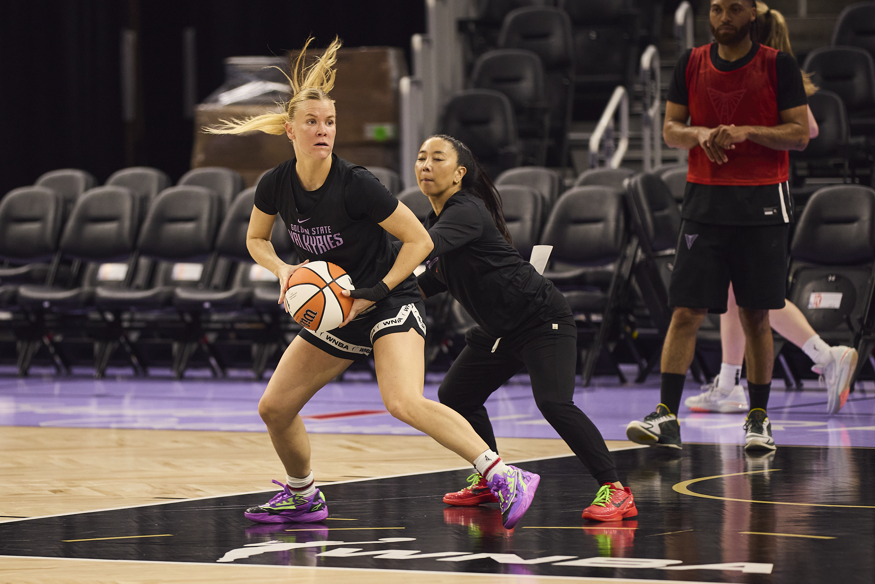 A basketball player in a &quot;Golden State Valkyries&quot; shirt is intensely dribbling with a defender close by. Others watch from the background on an indoor court.