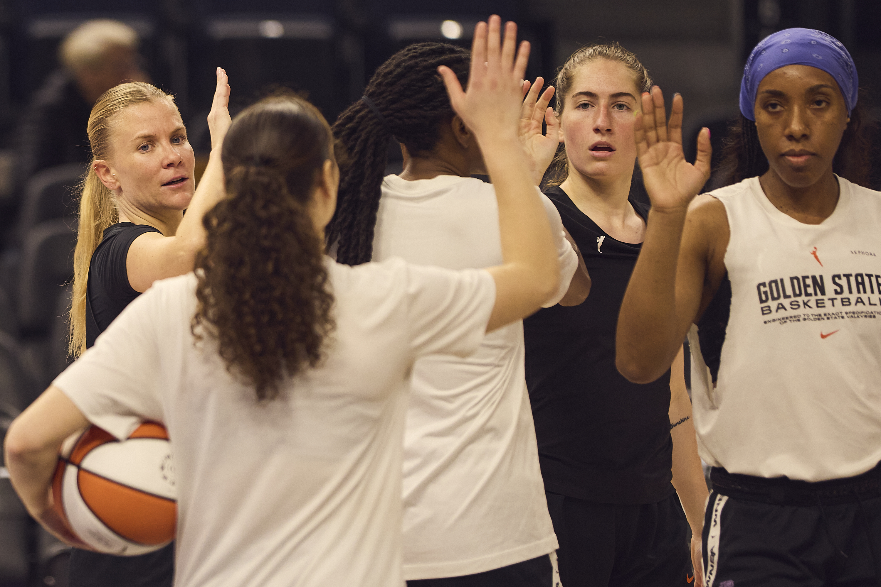A group of female basketball players, wearing practice attire, are engaged in a high-five gesture, suggesting camaraderie and teamwork on a basketball court.