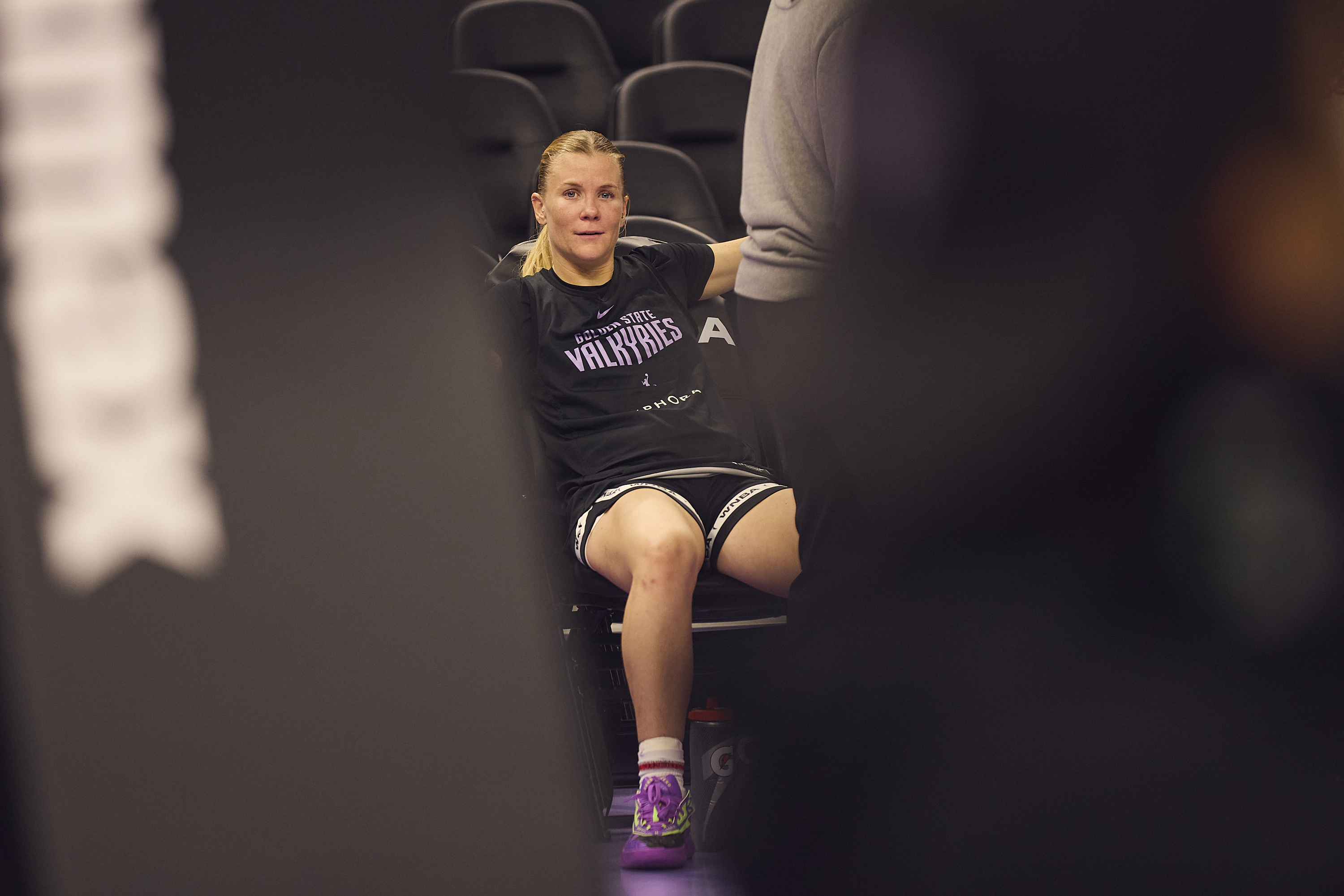 A woman in sportswear sits on a bench with her arms resting, wearing a &quot;Valkyries&quot; shirt and purple shoes. The foreground is blurred, creating a focused view.