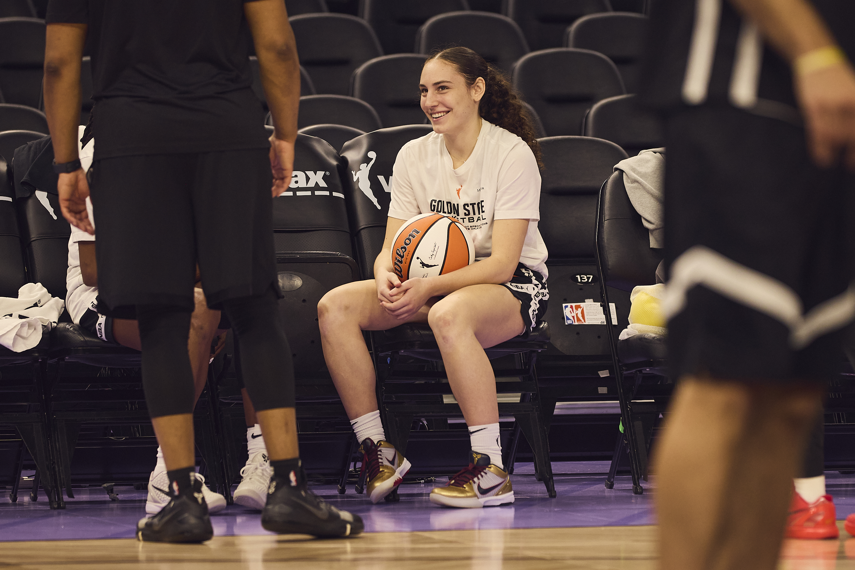 A person in a white shirt and shorts sits on a bench holding a basketball, smiling. Others stand nearby, wearing athletic attire, on a basketball court.