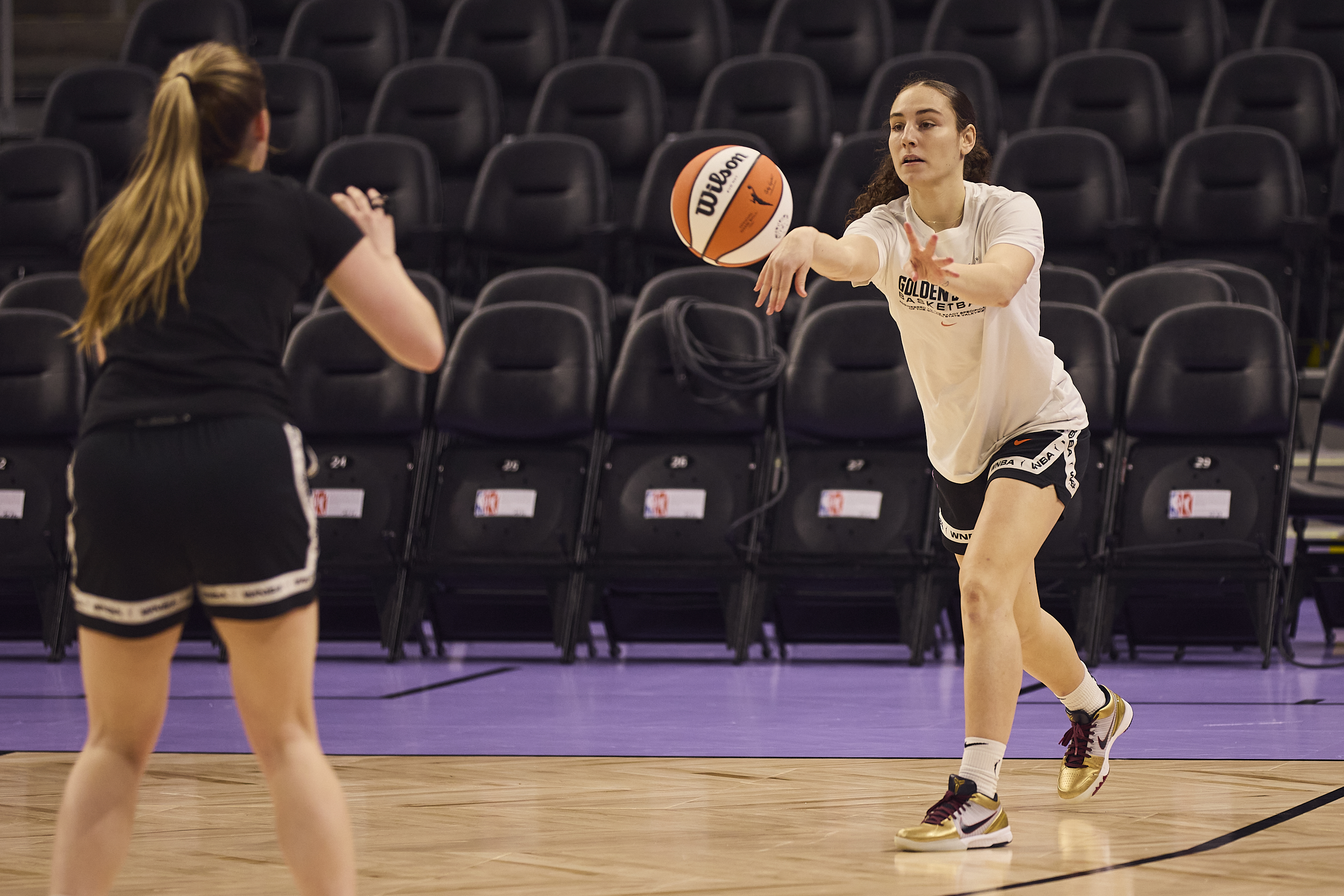 Two women are on a basketball court, one passing an orange-and-white ball. They're wearing athletic clothes, and empty seats are behind them.