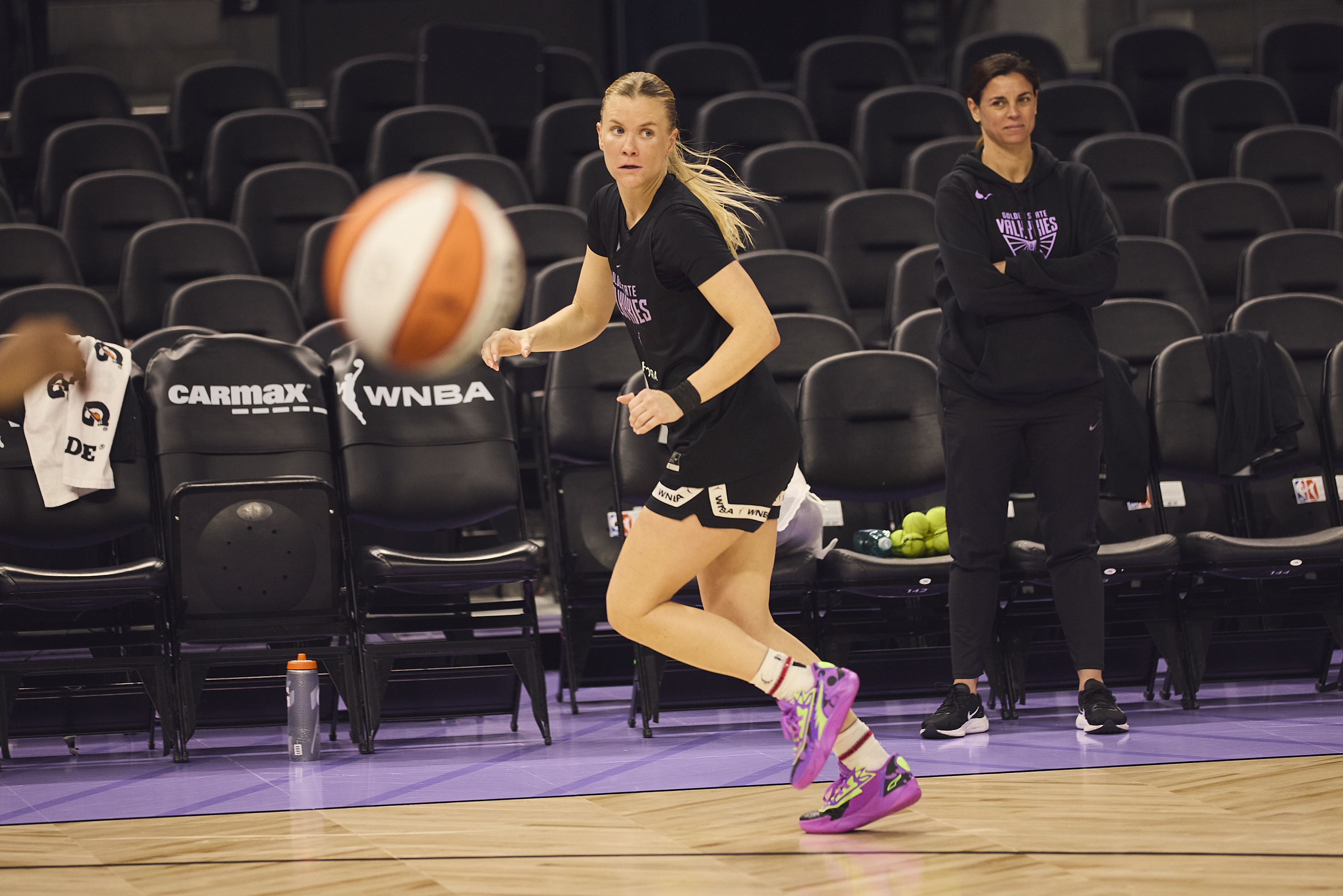 A woman in athletic gear is mid-jump with a basketball nearby. Another person watches from the sidelines. Empty chairs and sports equipment are visible.