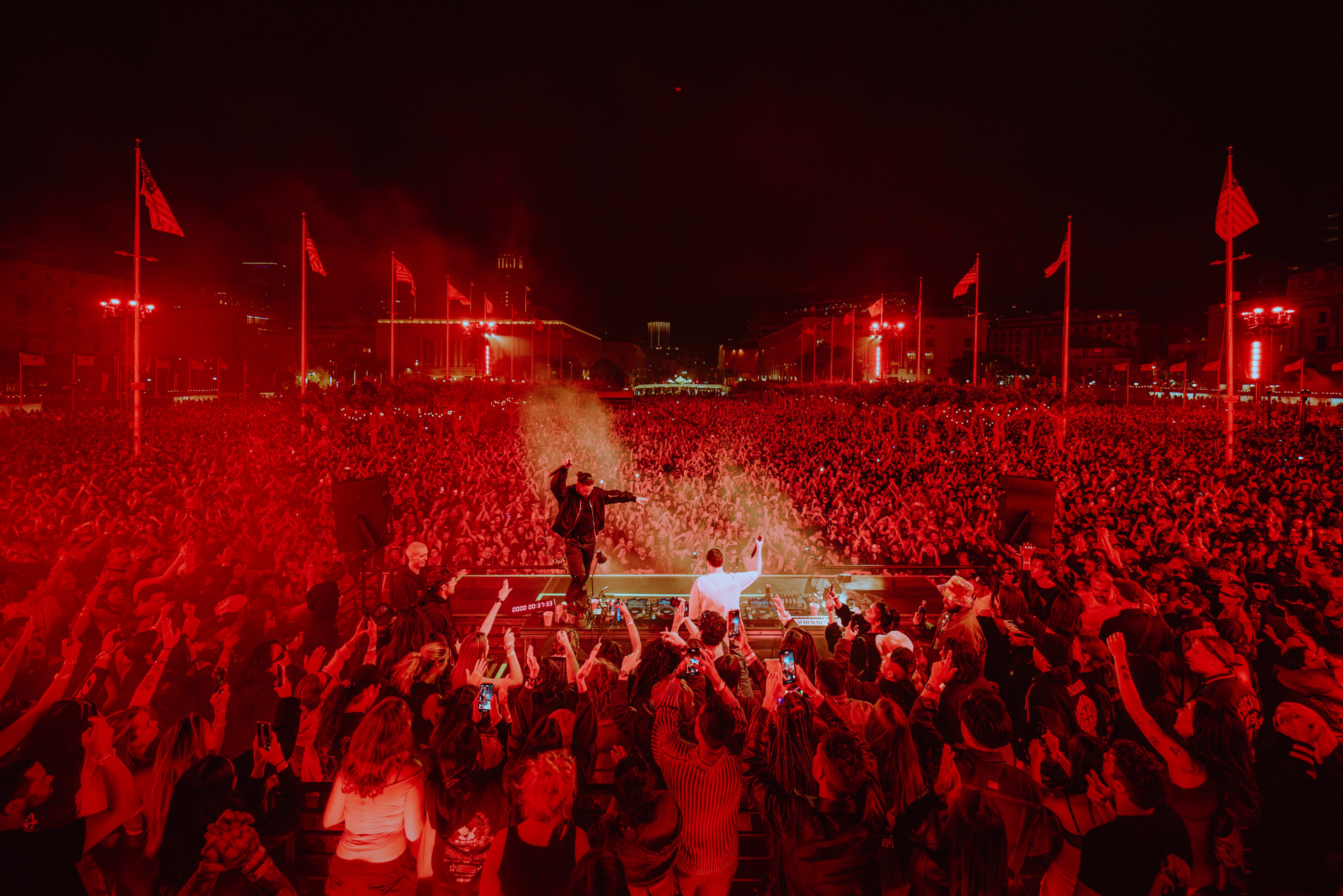 A musician performs on stage before a massive, energetic crowd at night, illuminated by red lighting and surrounded by waving flags.