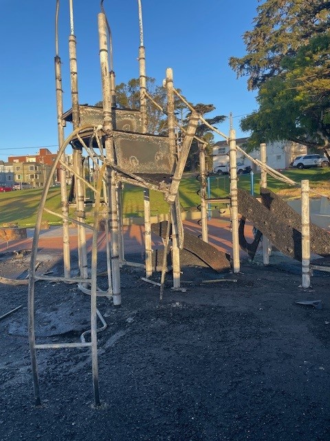 The image shows a burned playground structure, with charred and melted components. The area is surrounded by grass and some buildings in the background.