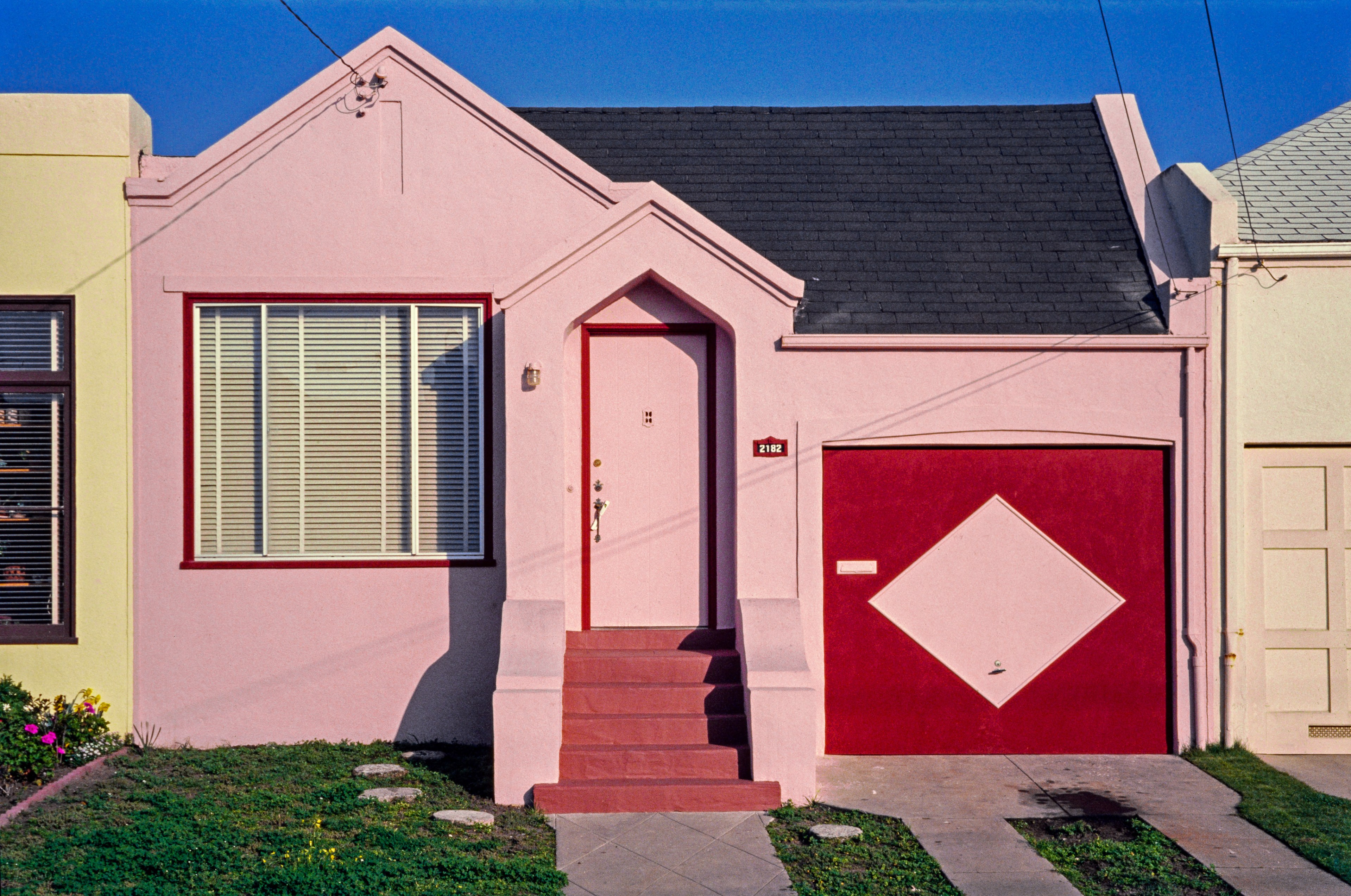 A small pink house with a red door and a garage featuring a diamond pattern. It has a neat lawn with stepping stones and a small flowerbed.
