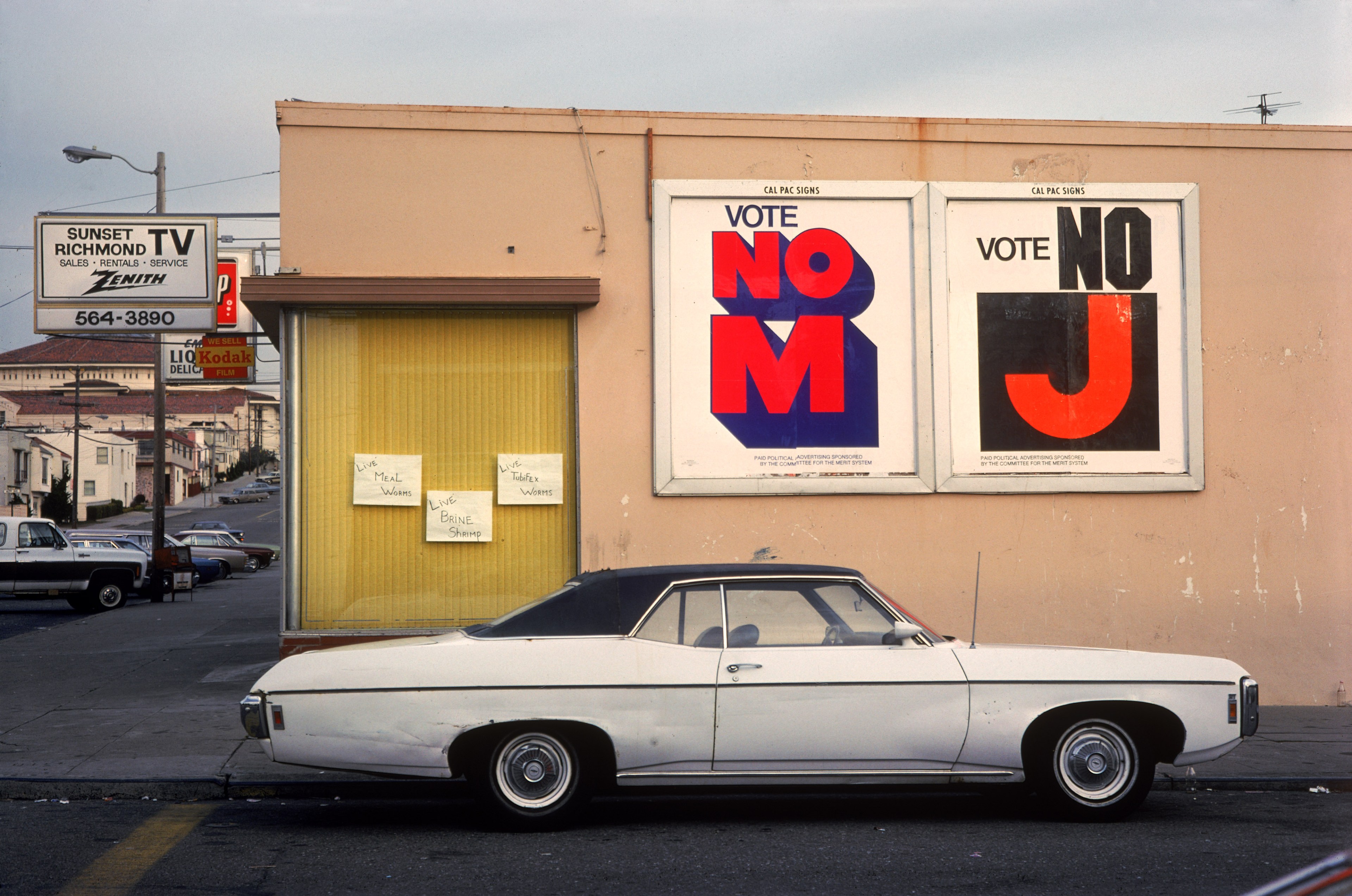 A white vintage car is parked in front of a building with posters saying &quot;VOTE NO M&quot; and &quot;VOTE NO J.&quot; A sign advertises live worms and shrimp.