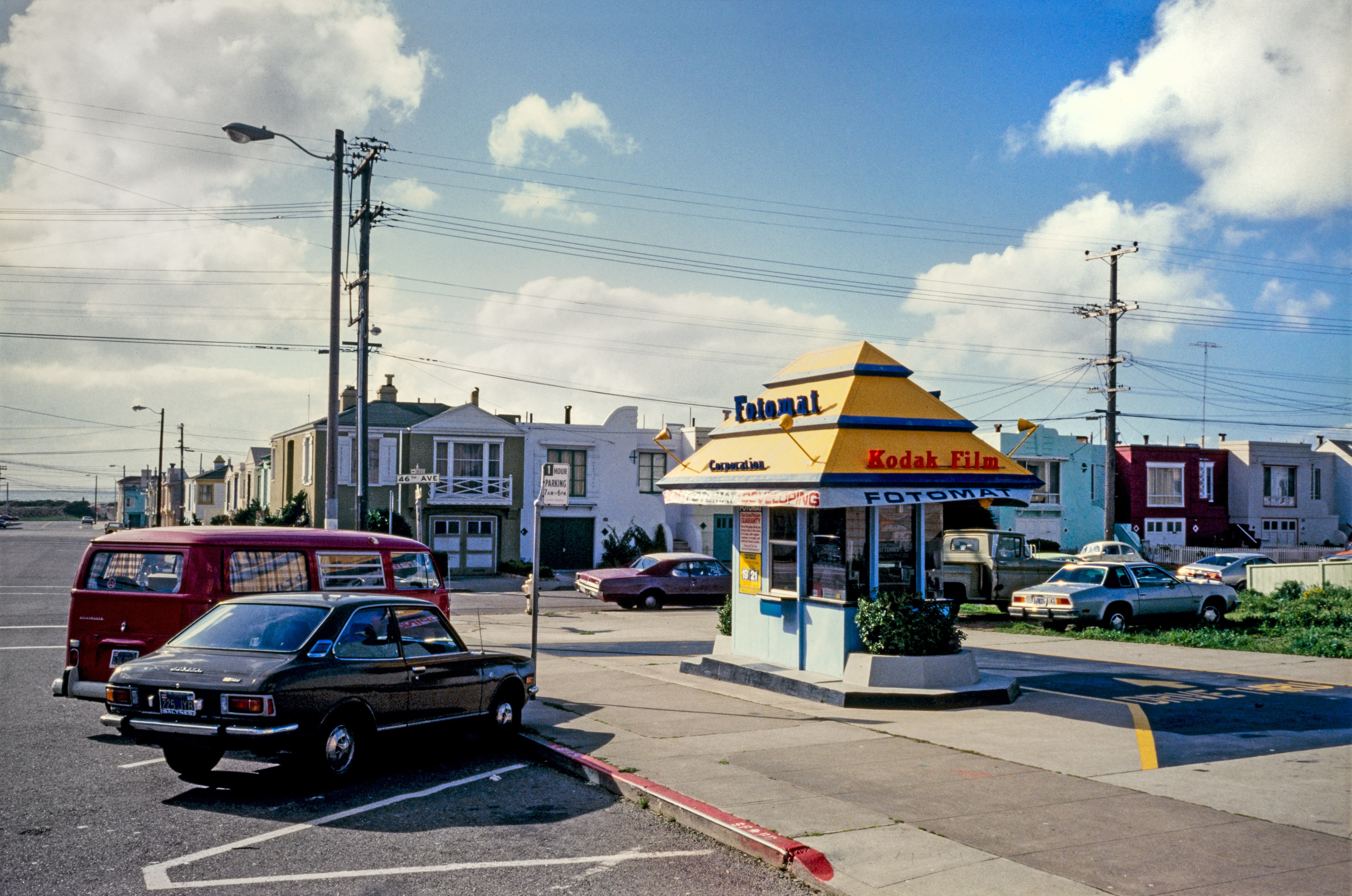 The image shows a small, yellow Fotomat kiosk in a parking lot, surrounded by vintage cars under a partly cloudy sky. Nearby are residential buildings and power lines.