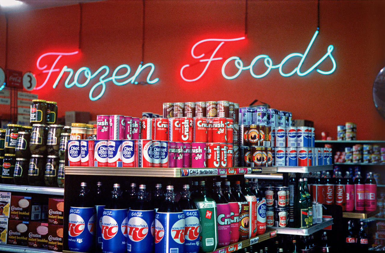 A grocery store aisle displays cans and bottles of soda, with brands like Diet Rite, Crush, and RC Cola. A neon sign above reads &quot;Frozen Foods.&quot;