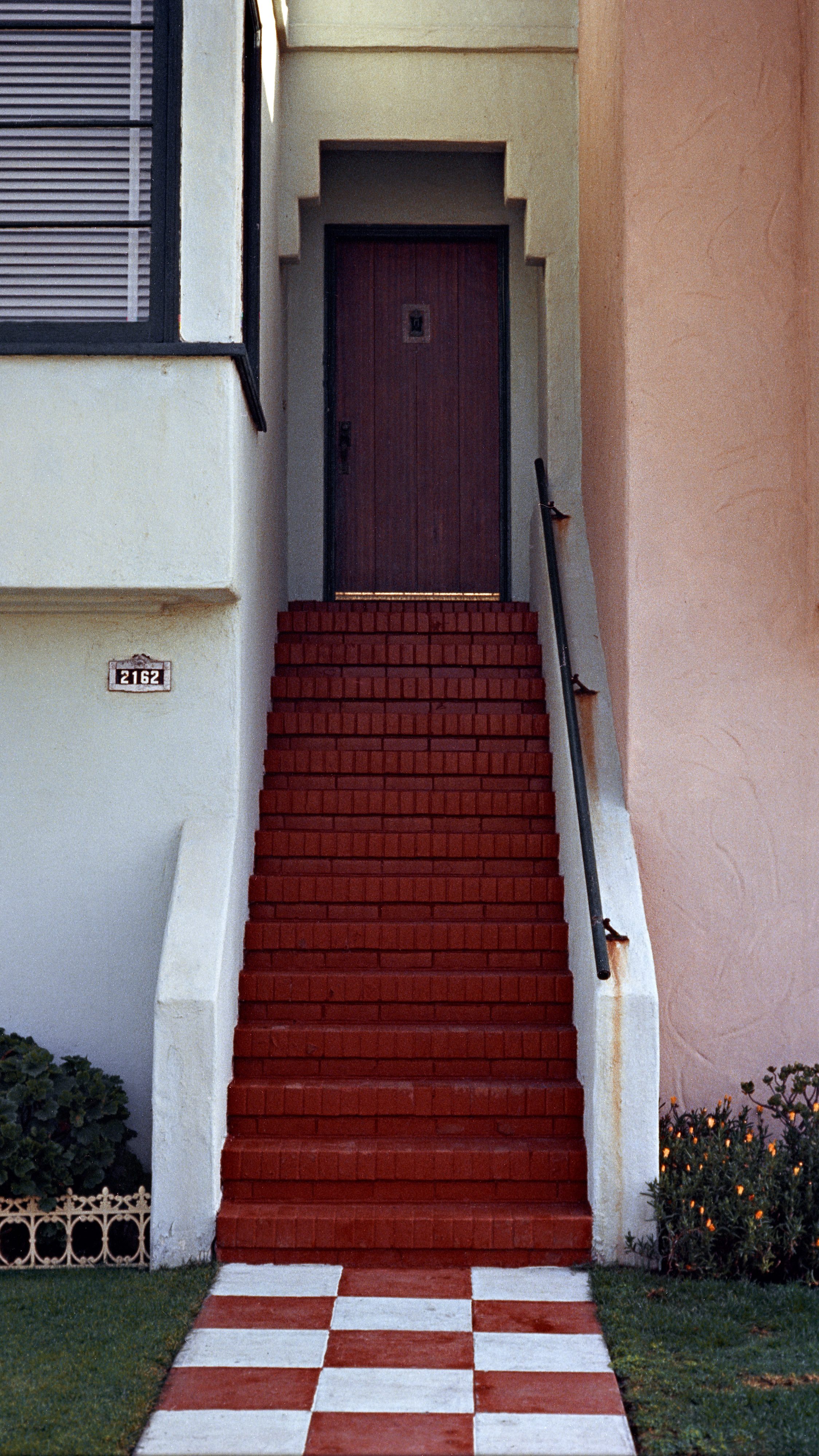 A red brick staircase leads up to a wooden door set in an arched entrance. The pathway features a red-and-white checker pattern. Plants flank the stairs.