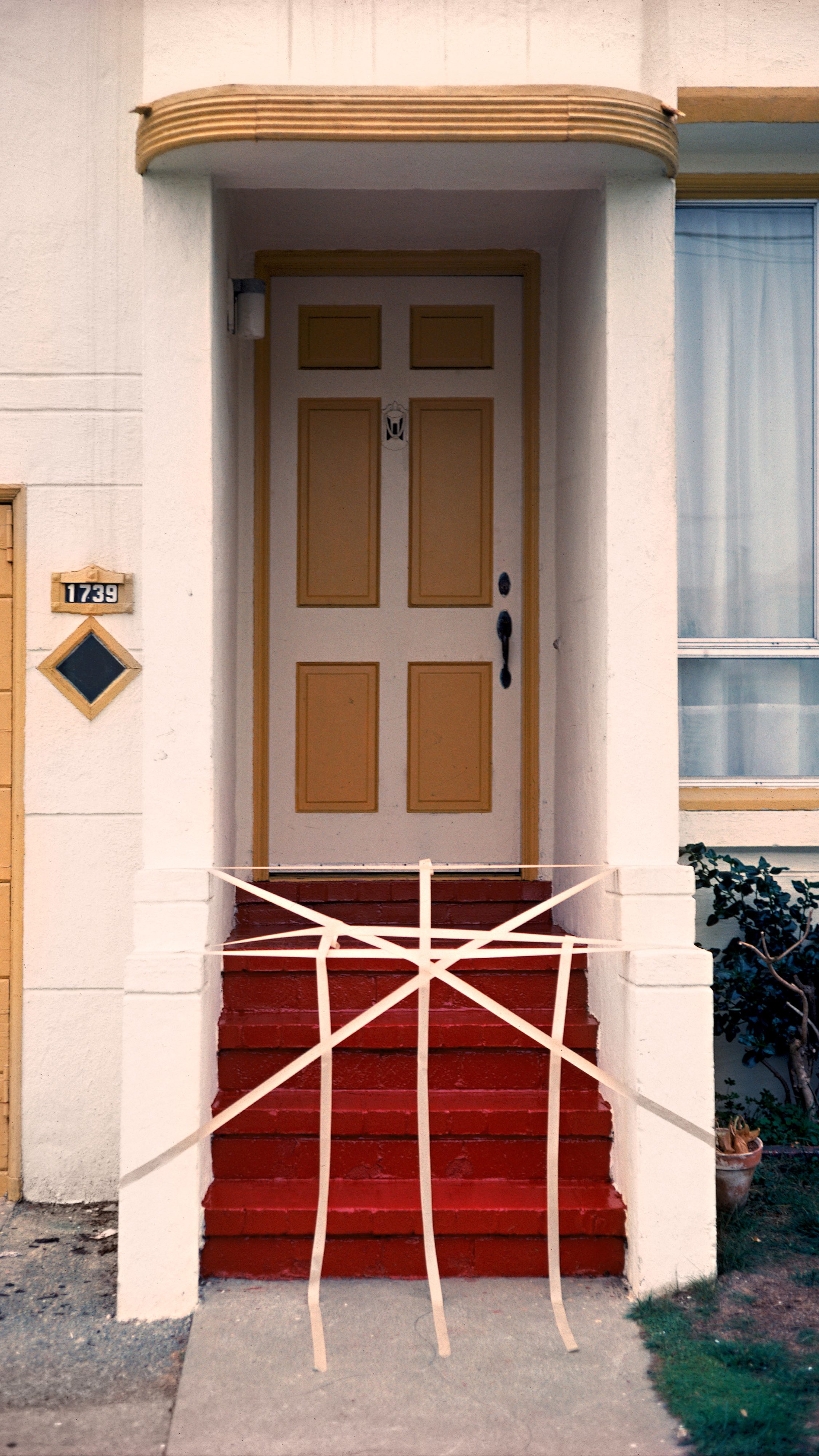 A red staircase leads to a tan, panelled door with white columns. The entrance is barricaded with white tape arranged in a web pattern.
