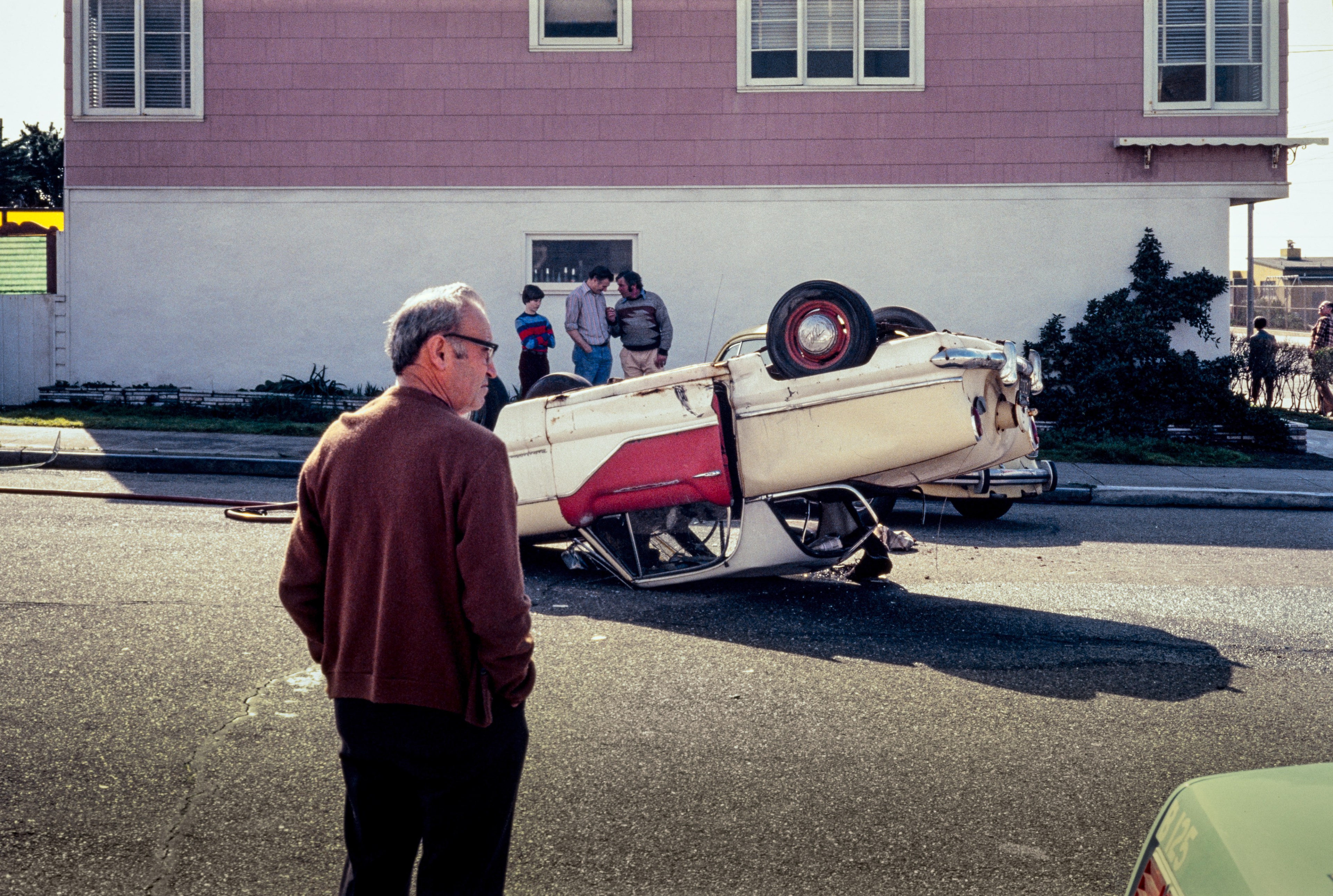 A man observes an overturned vintage car on a street. A small group of people gathers nearby, and a pink house with a white lower half is in the background.