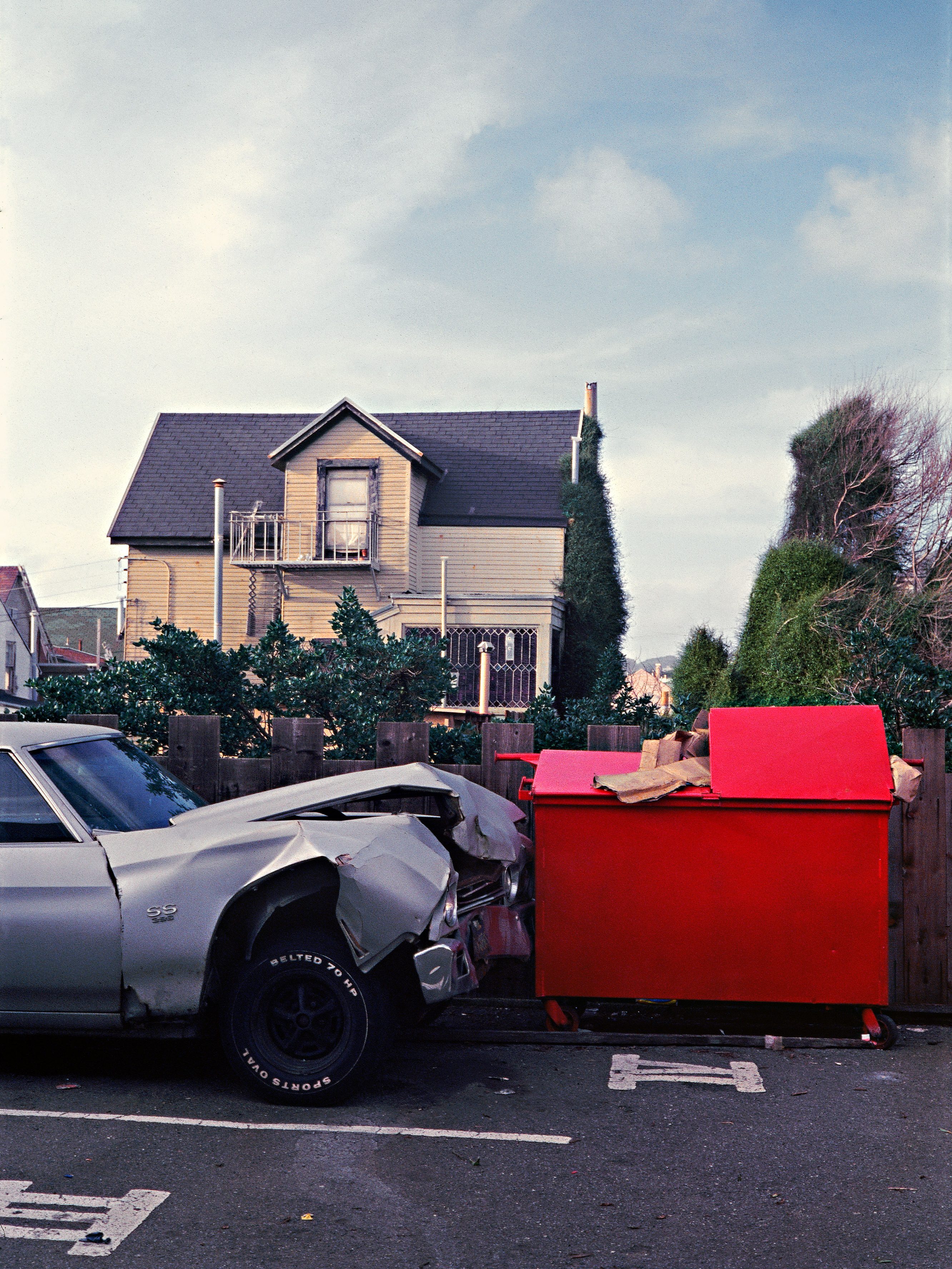 A damaged silver car is hitched to a bright red dumpster in a parking lot. Behind is a two-story house with a peaked roof, surrounded by trees and bushes.
