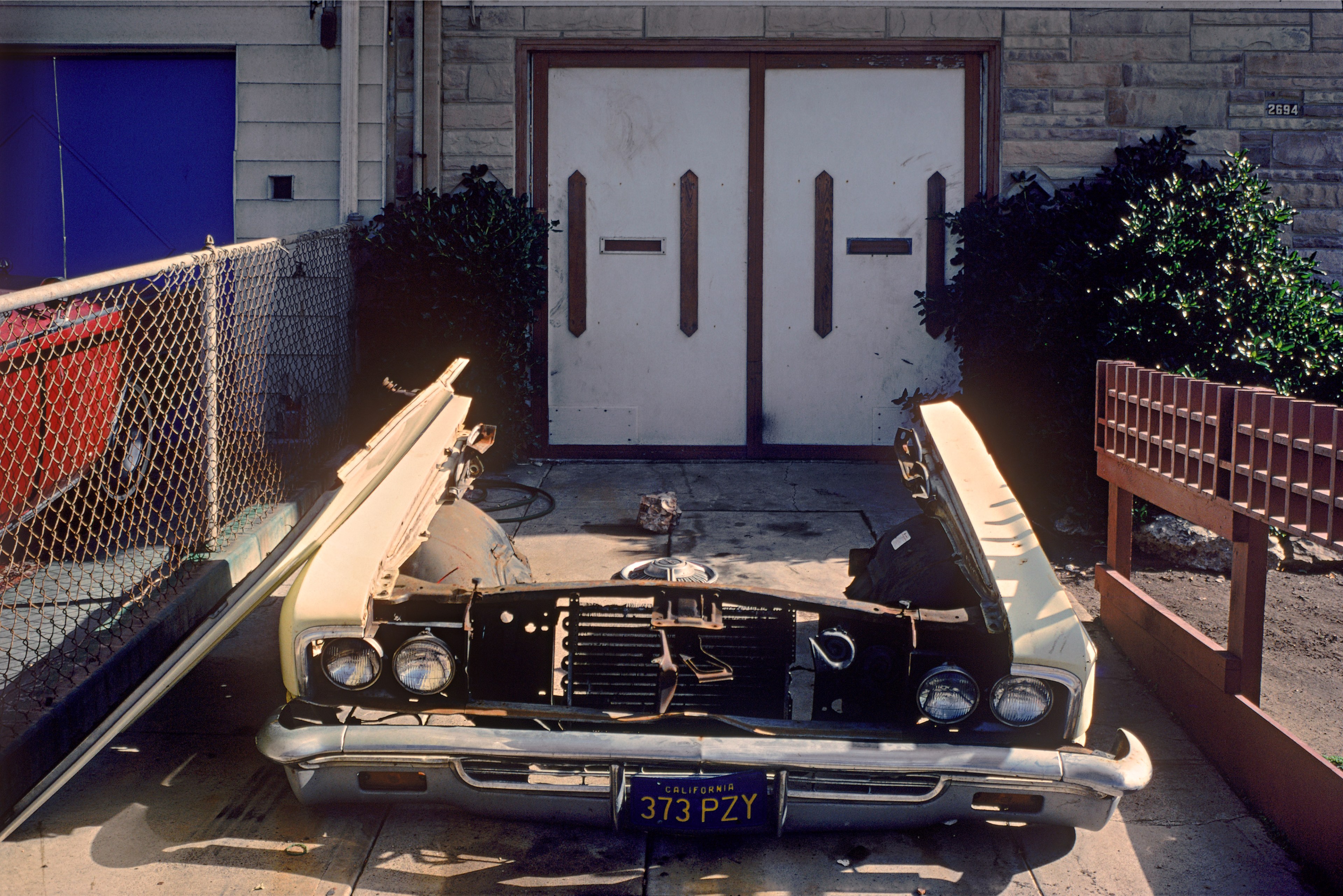 A dismantled car front is placed in a driveway, framed by a chain-link fence on the left and a wooden fence on the right, in front of double garage doors.