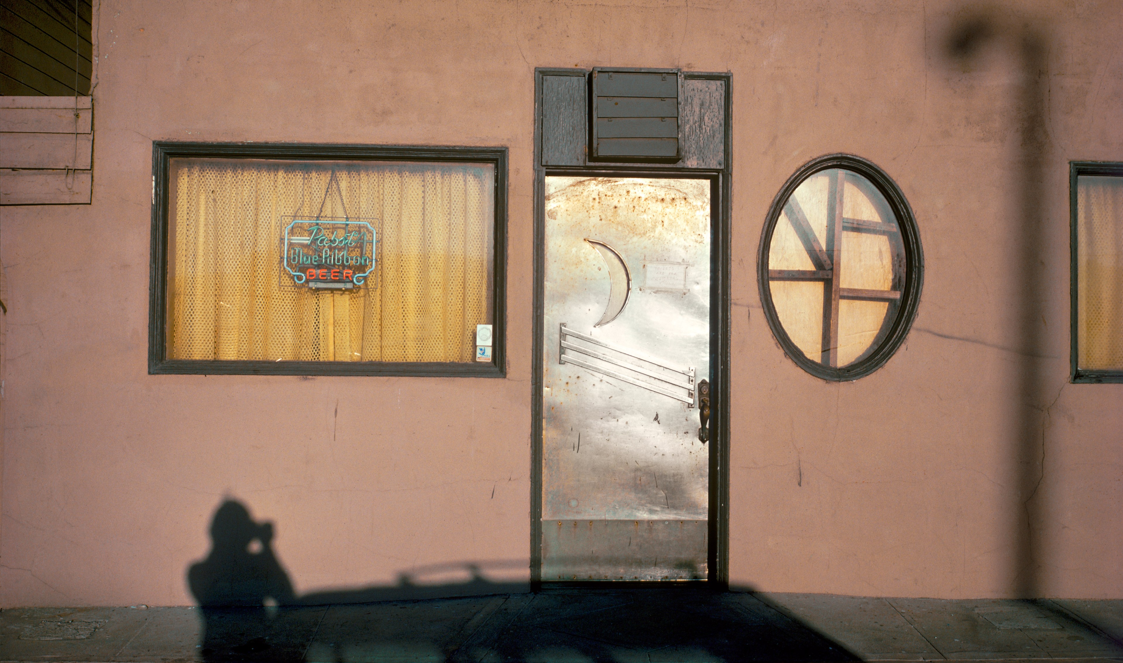 A weathered building facade with a metal door featuring a moon shape, a curtain-covered window with a beer sign, and a shadow of a person holding a camera.
