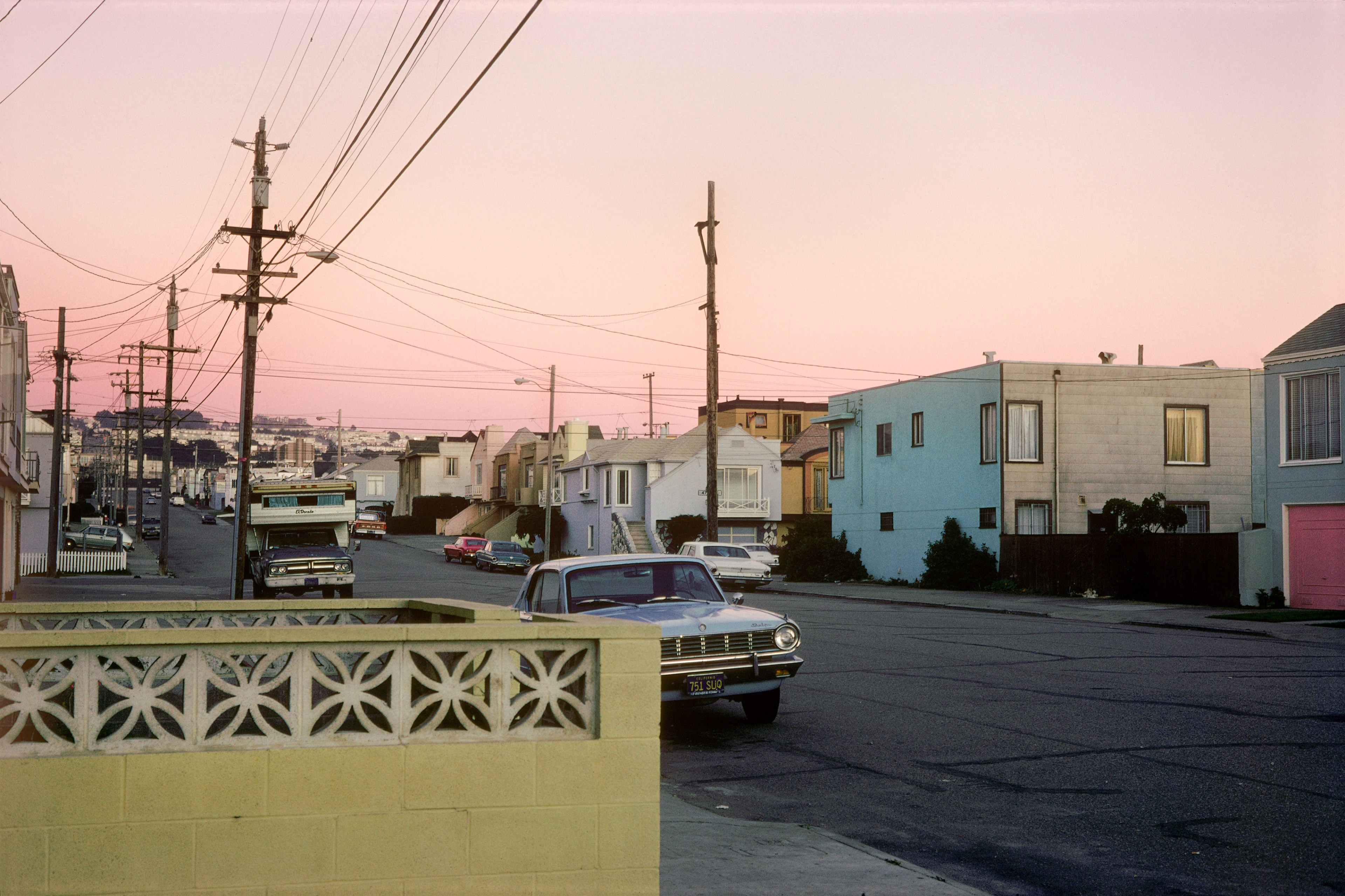 The image shows a quiet street at sunset with a pink sky, lined by pastel-colored houses. A 60s-style car and RV are parked along the road.
