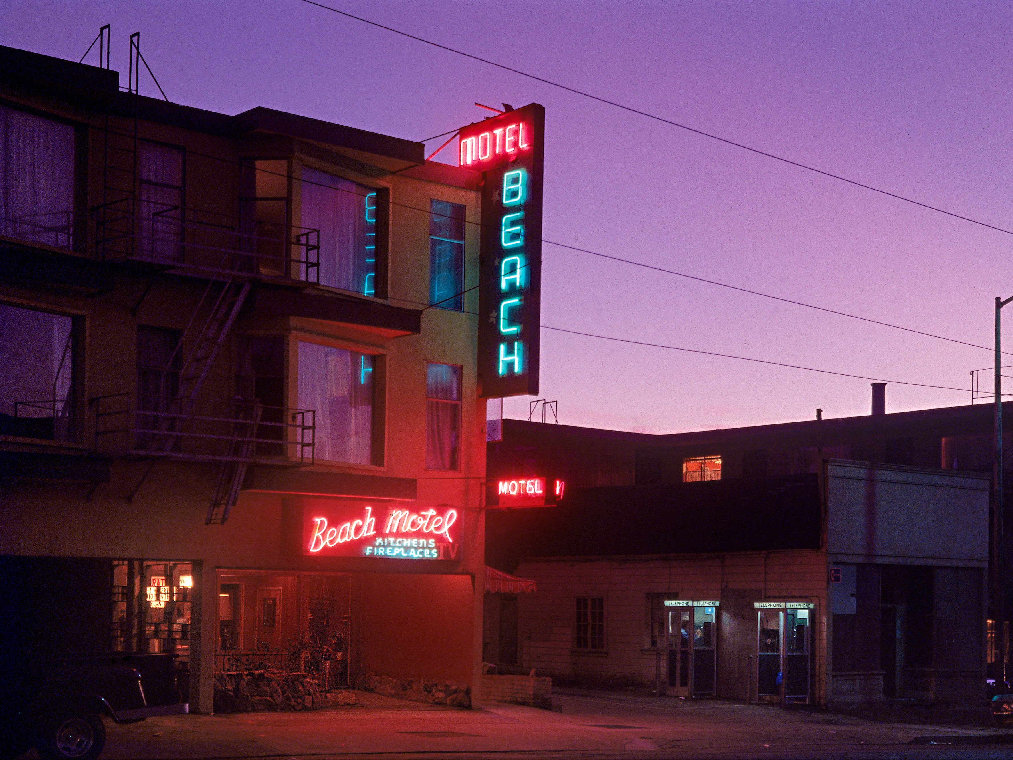The image shows a vintage motel with neon signs reading &quot;Beach Motel,&quot; &quot;Kitchens,&quot; and &quot;Fireplaces.&quot; It's dusk, casting a purple hue over the scene.