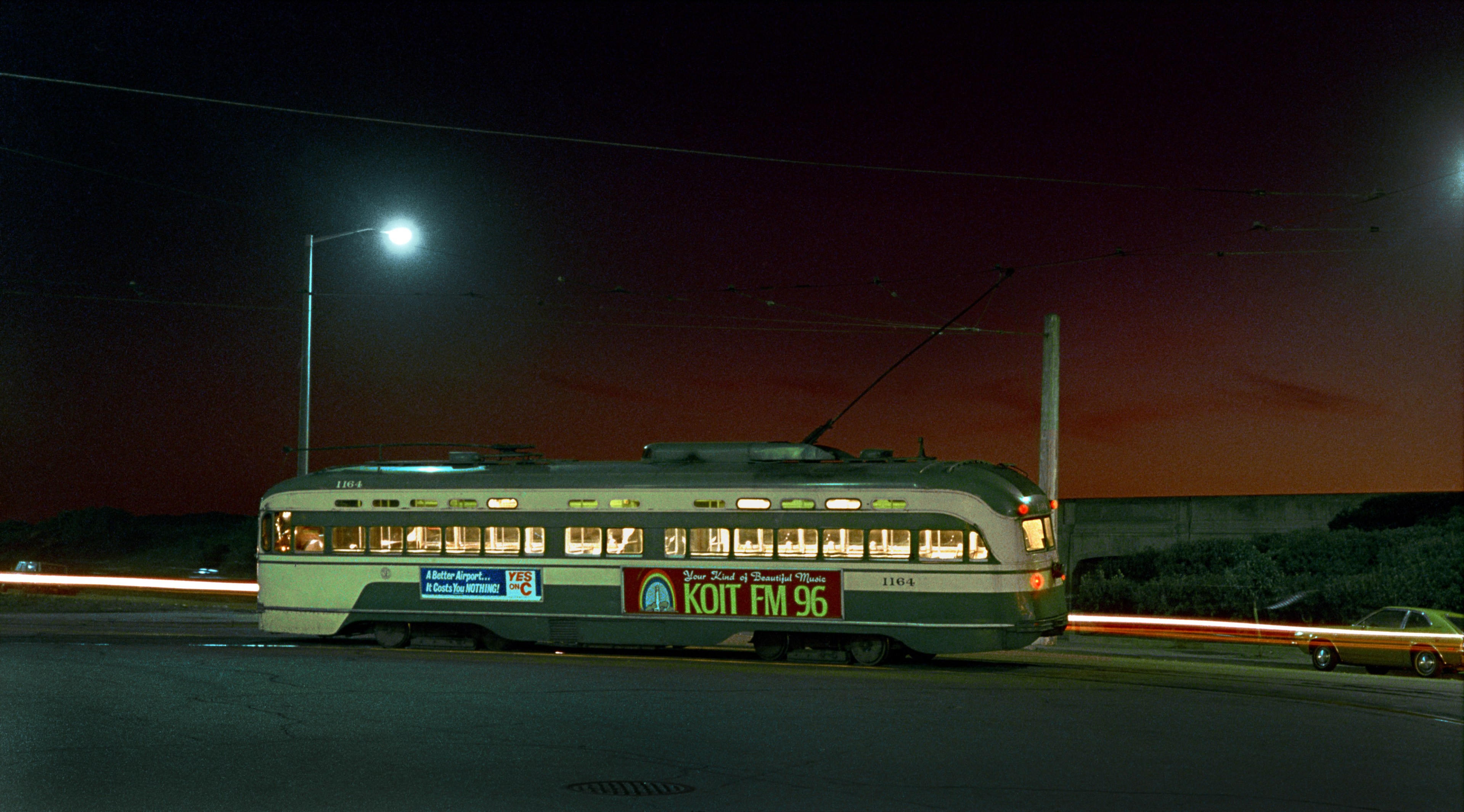 A vintage tram is illuminated at night under a streetlamp, with a vibrant red and orange sunset in the background and a passing car's light streaks.