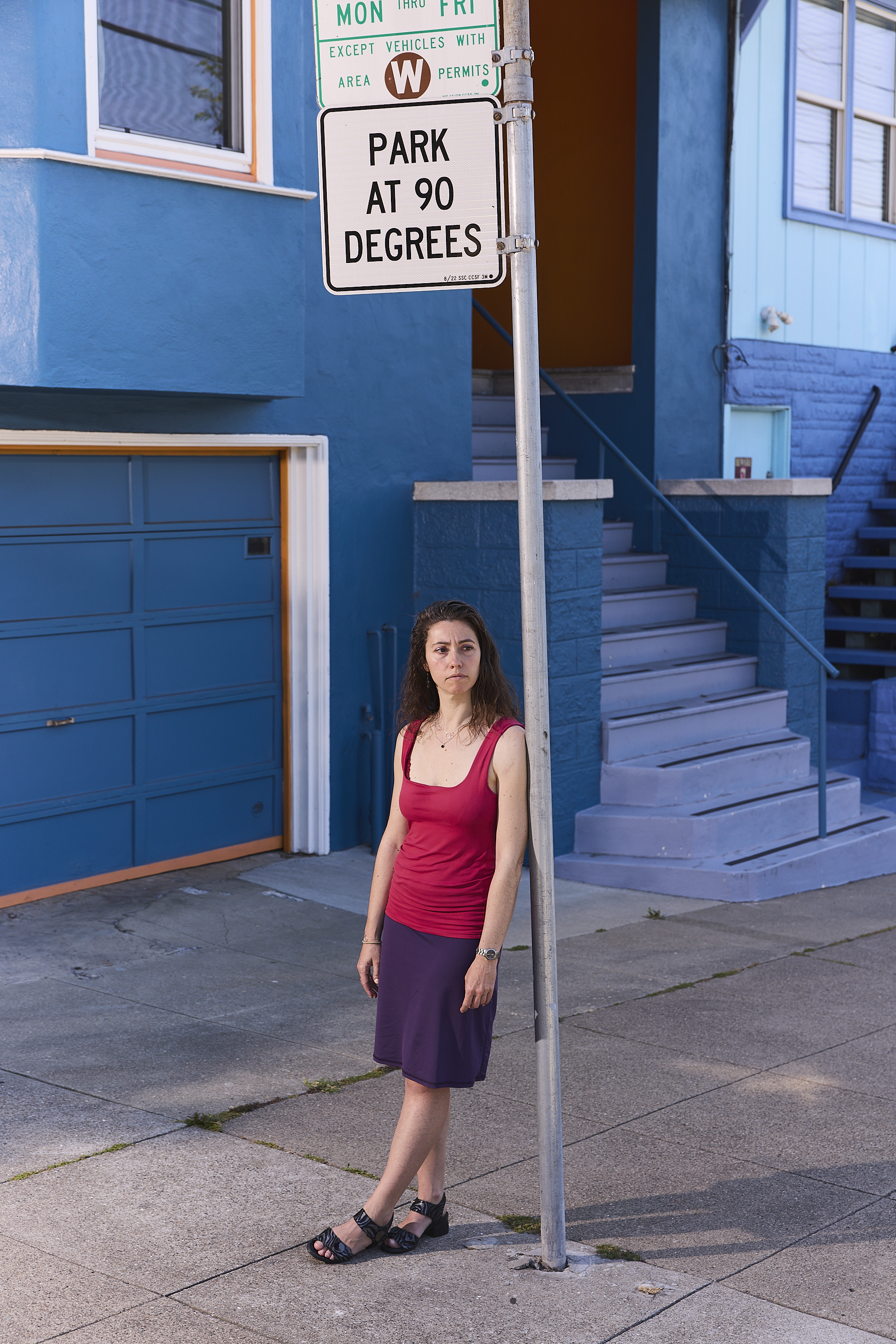 A woman in a red top and purple skirt stands next to a &quot;Park at 90 Degrees&quot; sign. Behind her is a blue building with a garage and stairs.