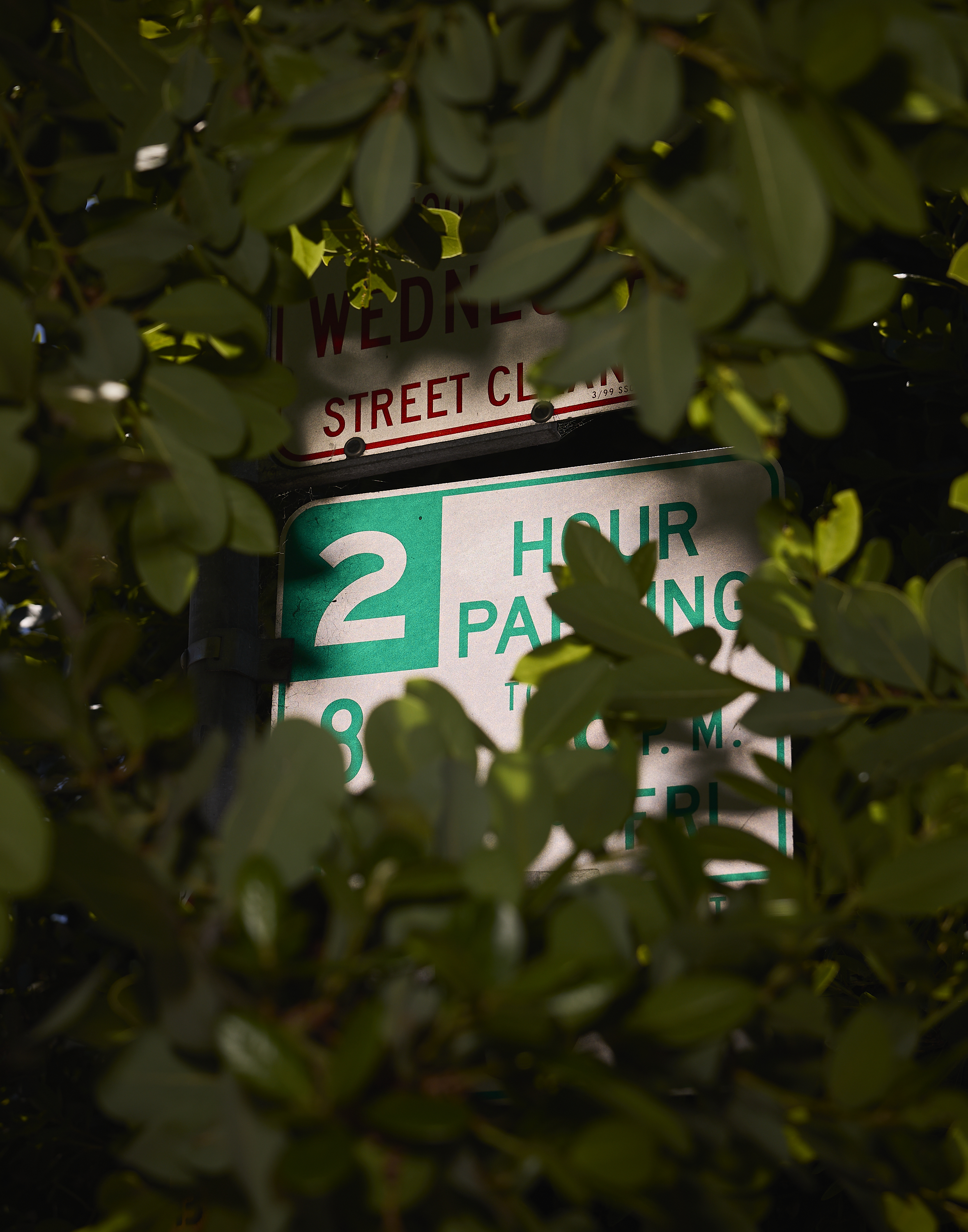 A parking sign is partially obscured by green leaves. The visible text indicates &quot;2 Hour Parking&quot; with some additional details not entirely clear.