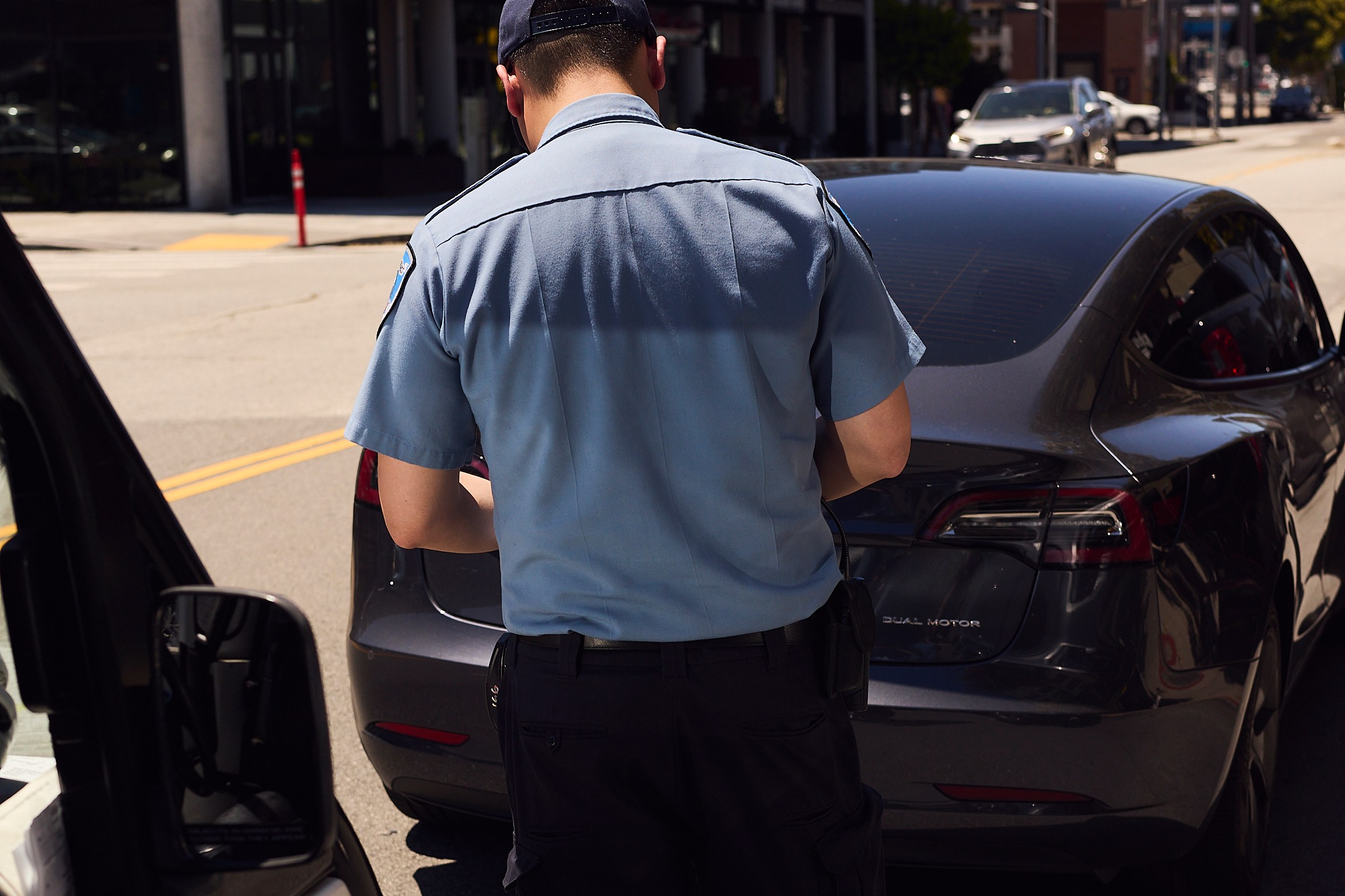 A uniformed officer stands on the street, writing a ticket beside a parked black car, with a building and other vehicles in the background.
