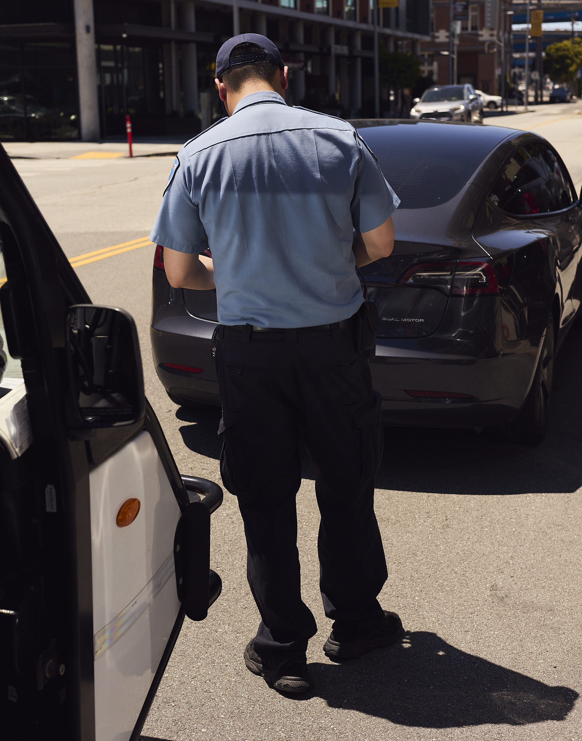 A uniformed officer stands on the street, writing a ticket beside a parked black car, with a building and other vehicles in the background.