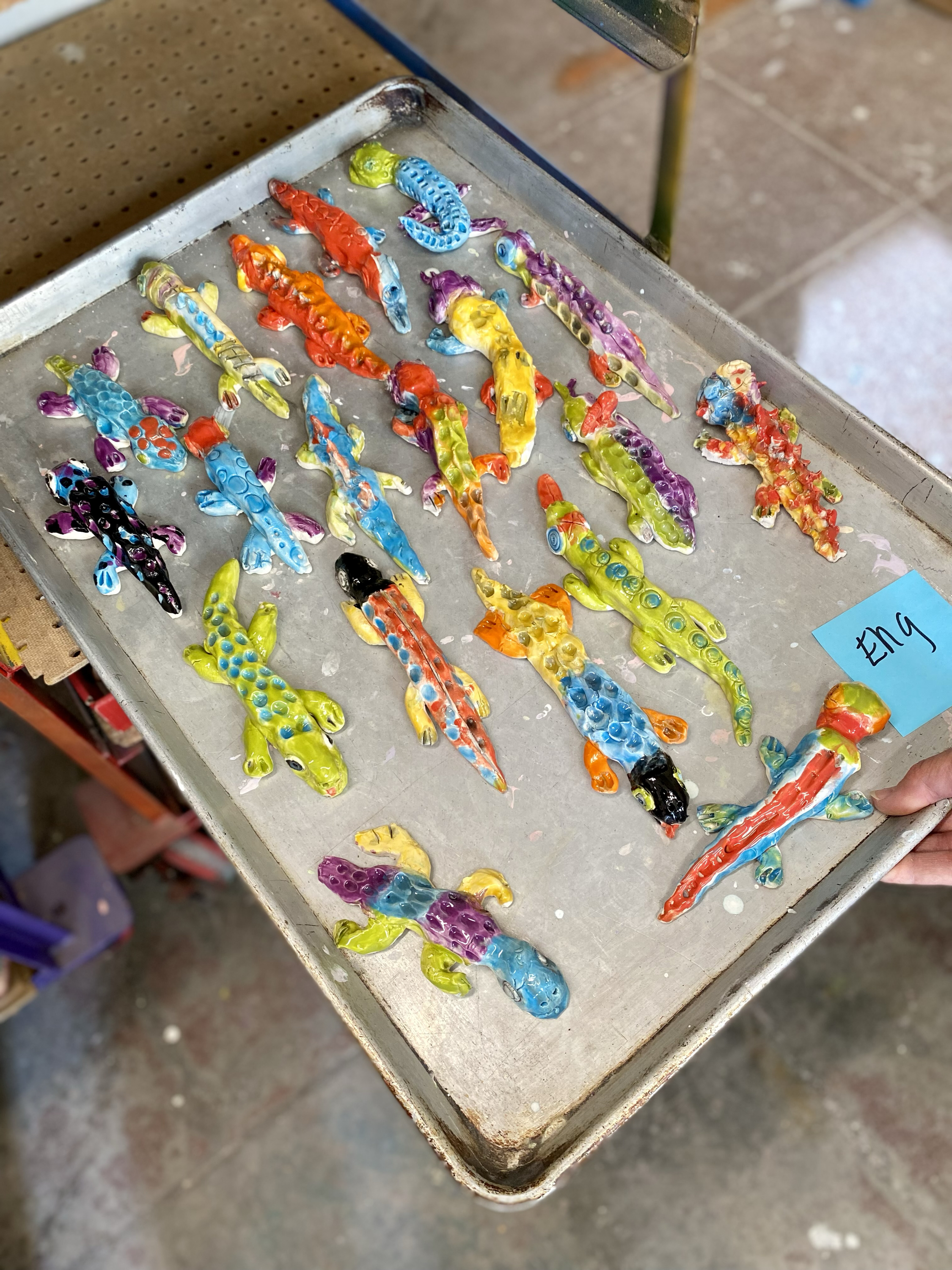 The image shows a metal tray filled with colorful, ceramic lizard figurines in various bright shades and patterns. A small blue paper is labeled &quot;E19.&quot;