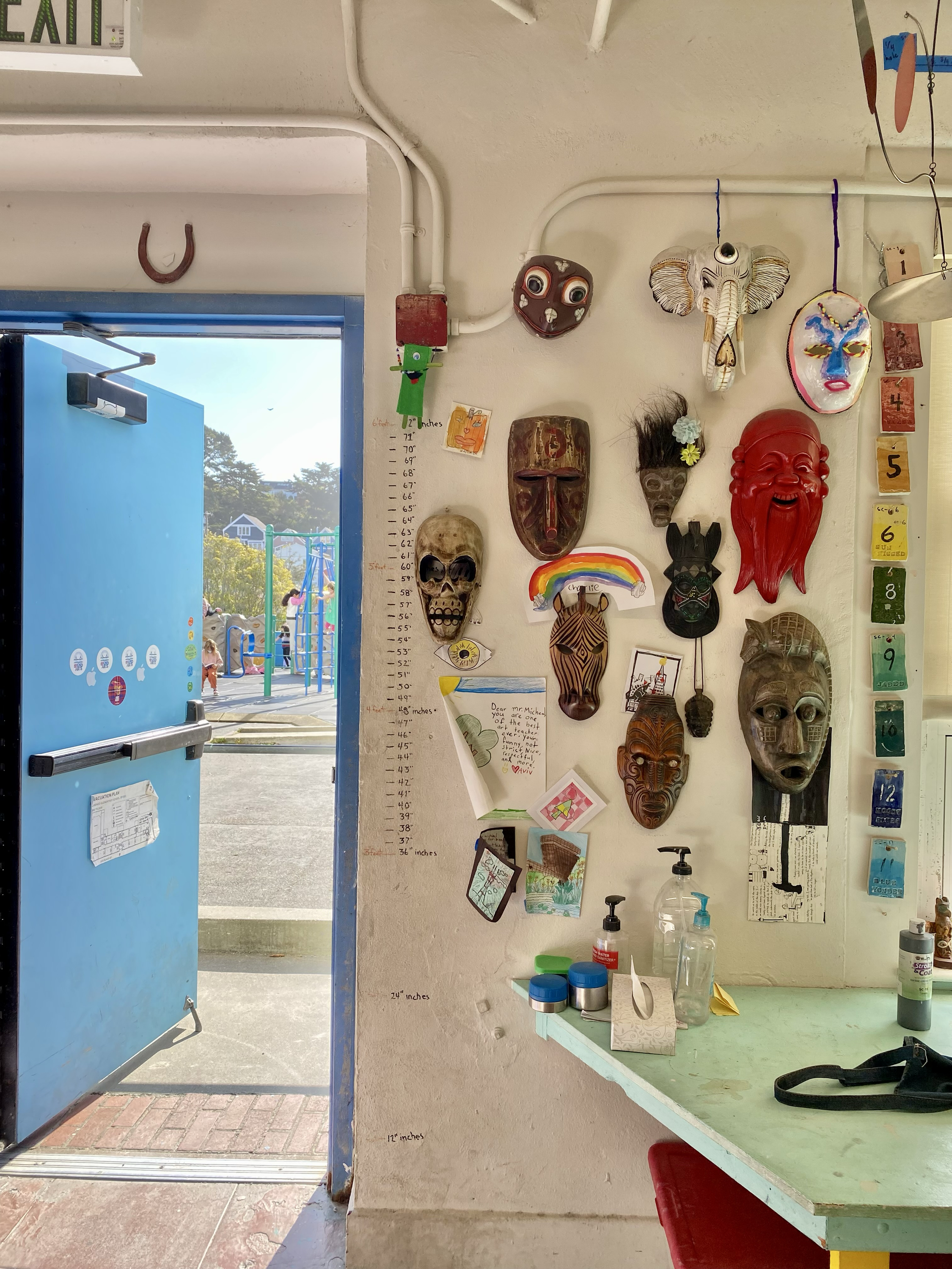 The image shows a wall decorated with various colorful masks and crafts beside an open blue door, revealing an outdoor playground. A table below holds art supplies.