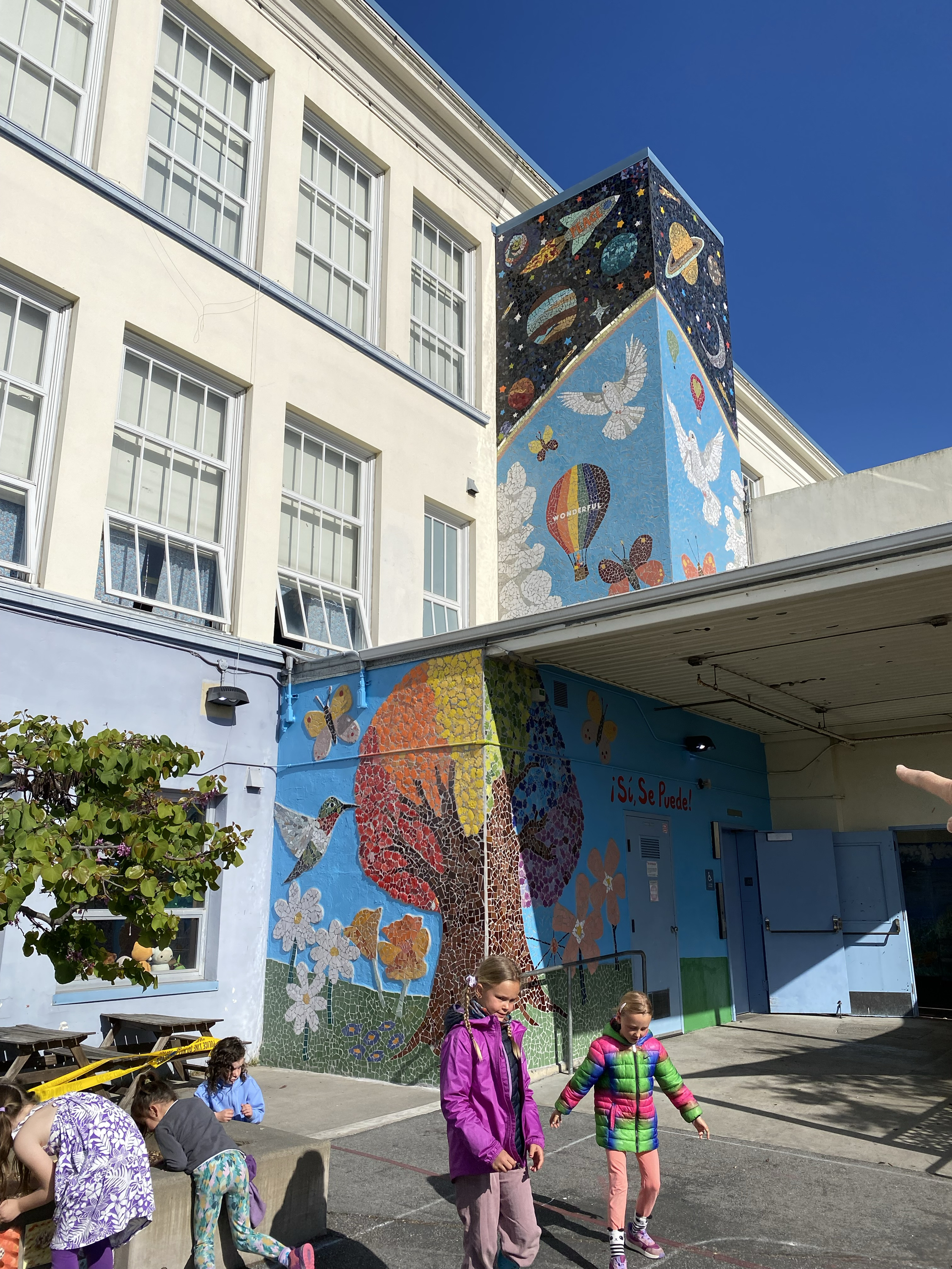 A colorful mural adorns a school building, featuring planets, doves, a hot air balloon, and a tree. Children play nearby under a clear blue sky.