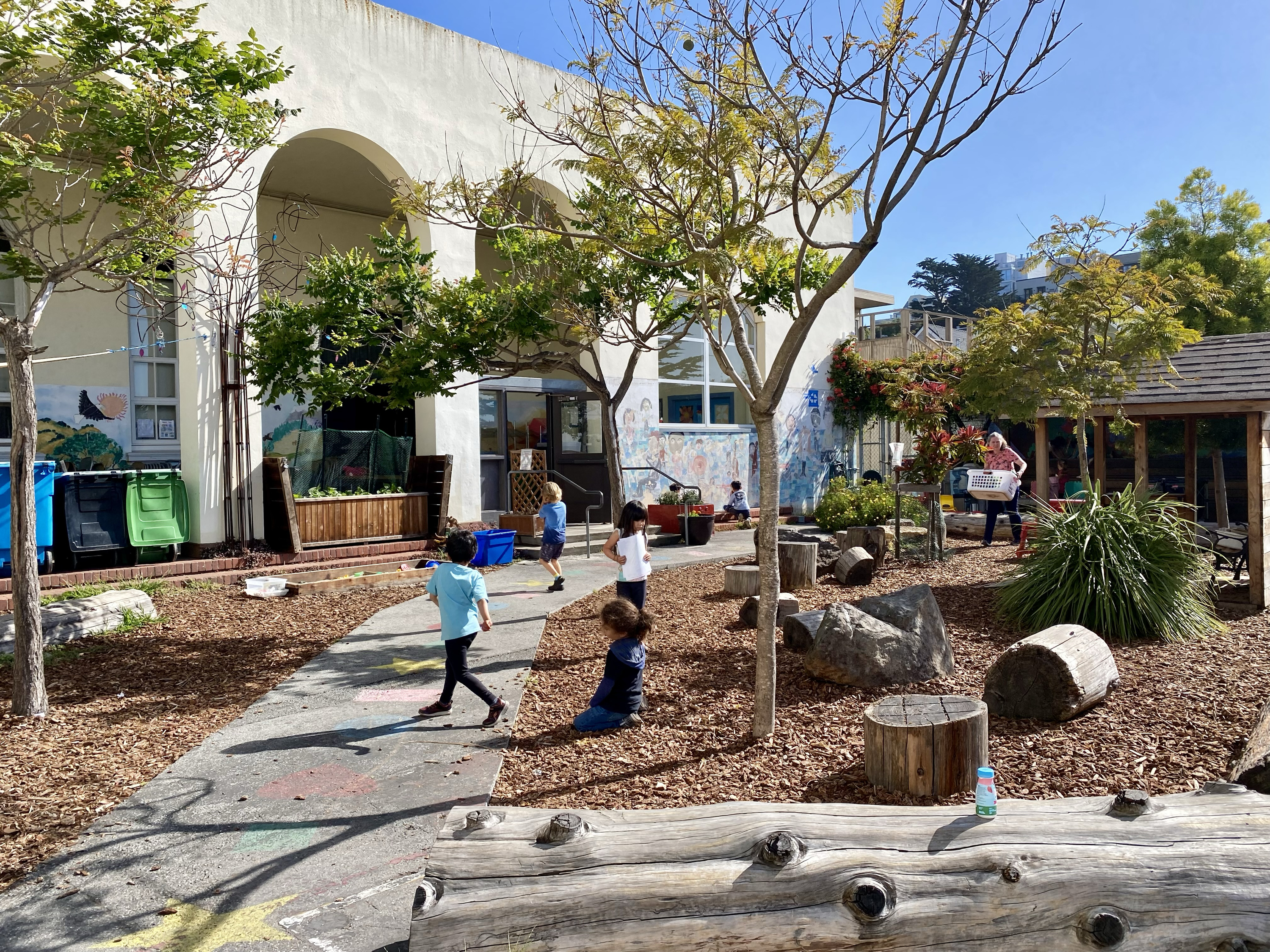 The image shows a sunny outdoor playground with children playing on a path amidst trees and logs, next to a building with colorful murals and recycling bins.
