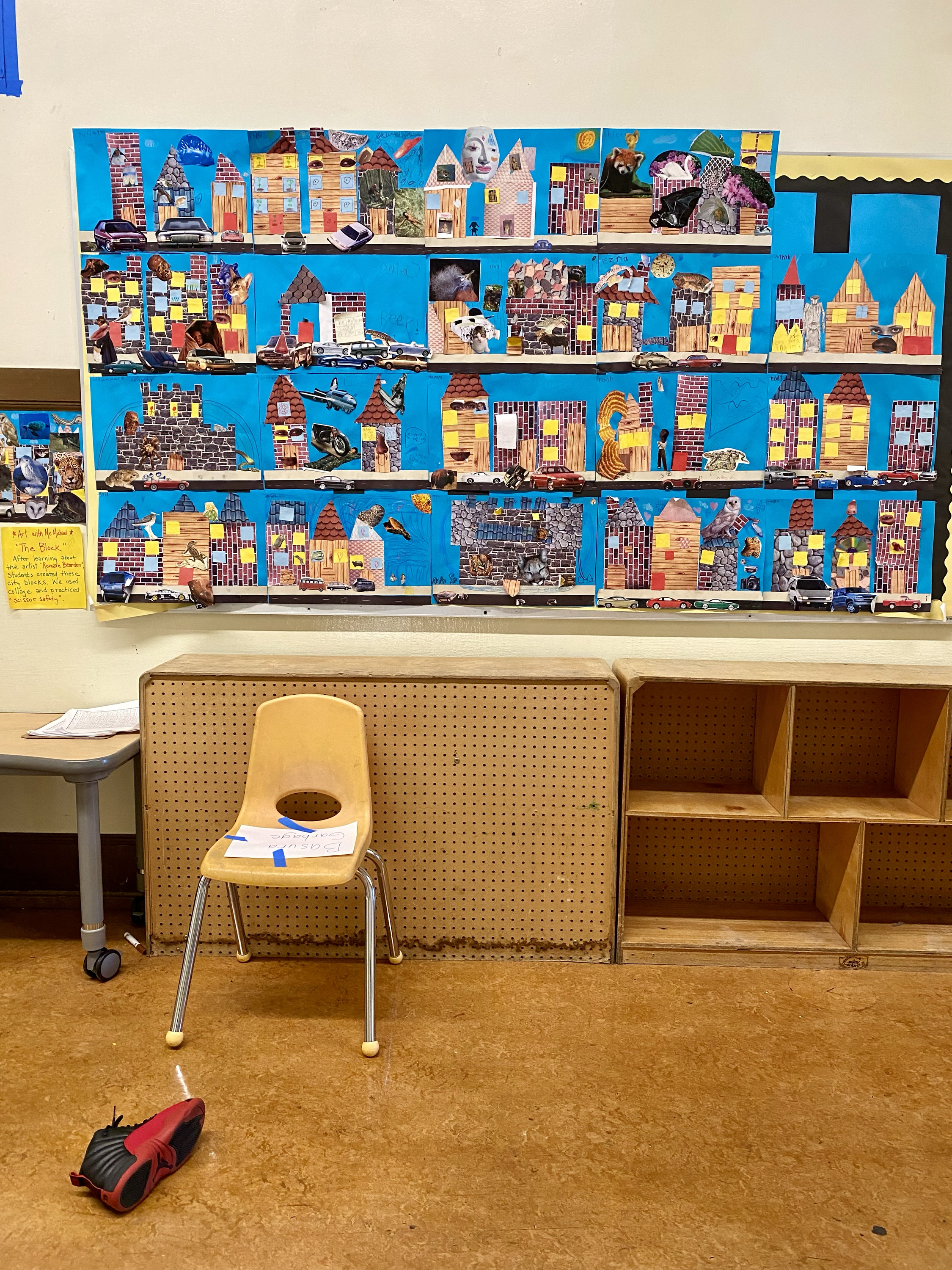 A colorful collage of houses on a blue background hangs on the wall. Below, a wooden chair holds a paper, and a red and black shoe lies on the brown floor.