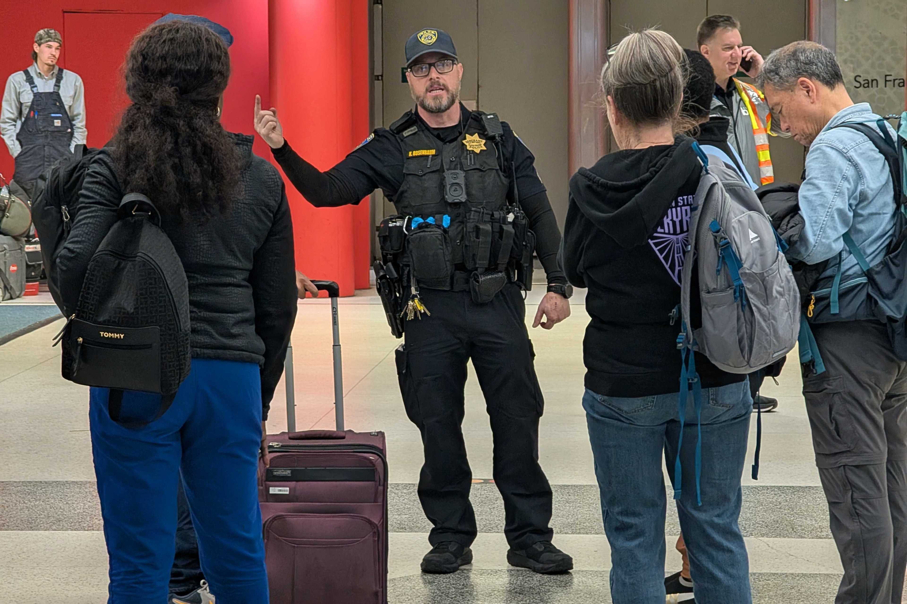 A police officer is speaking to a small group. One person has a suitcase and a backpack. Others are holding backpacks, and one uses a phone, with a red wall behind.