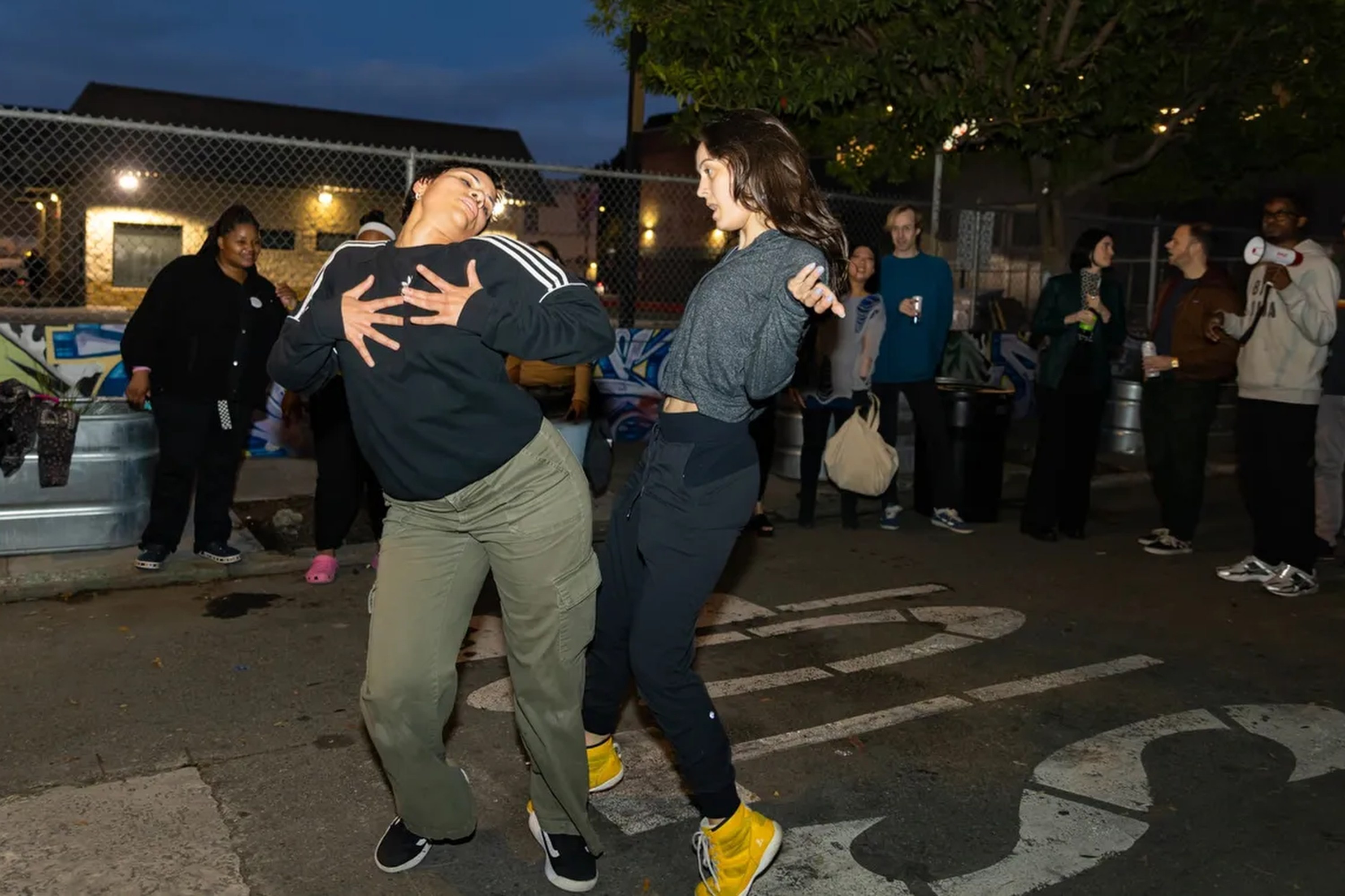 Two people are dancing energetically on a street at night, while a small crowd watches and some hold drinks. There's a chain-link fence and graffiti in the background.