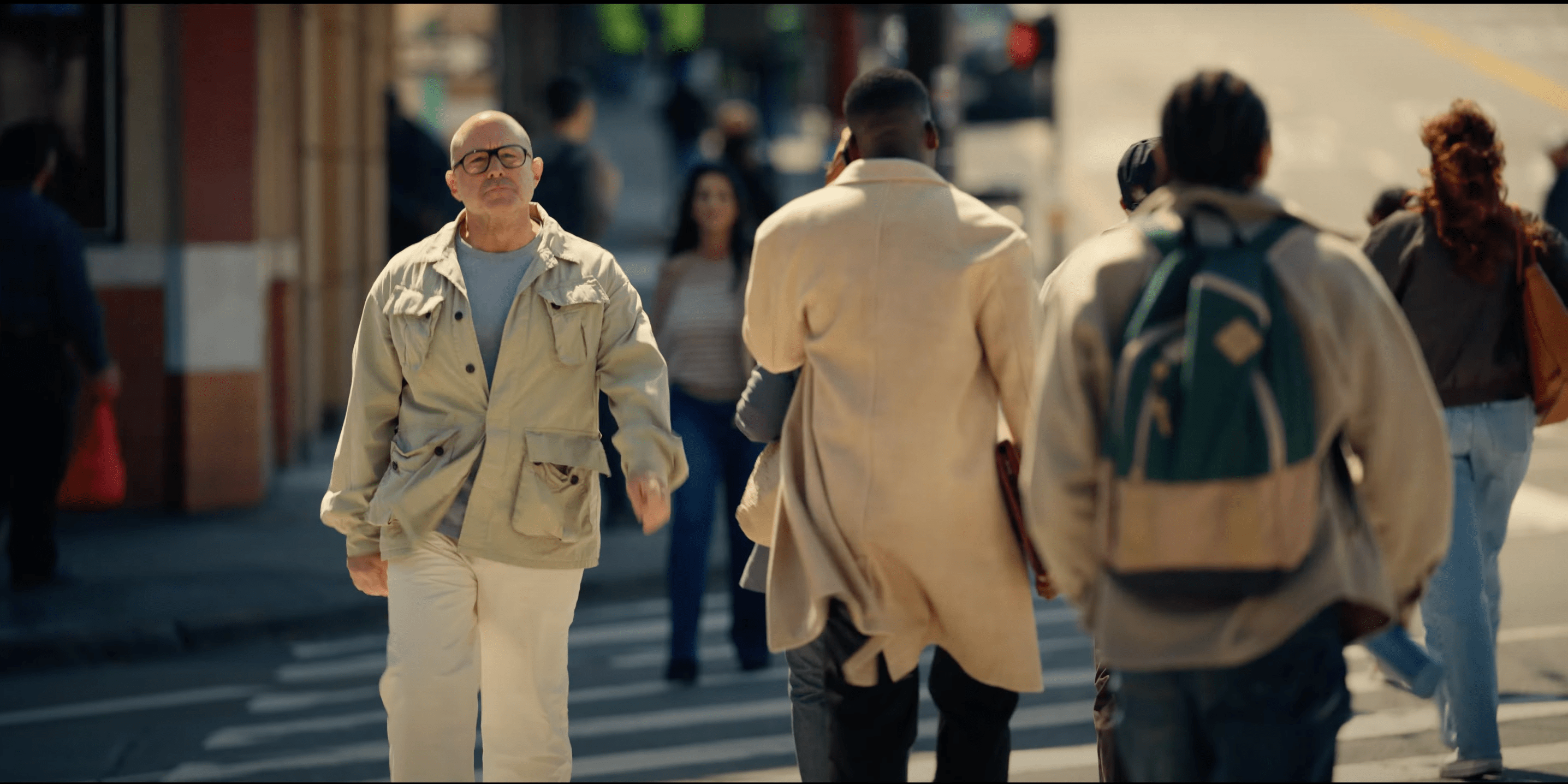 A man in a light jacket walks toward the camera on a busy sidewalk, surrounded by other people going in the opposite direction.