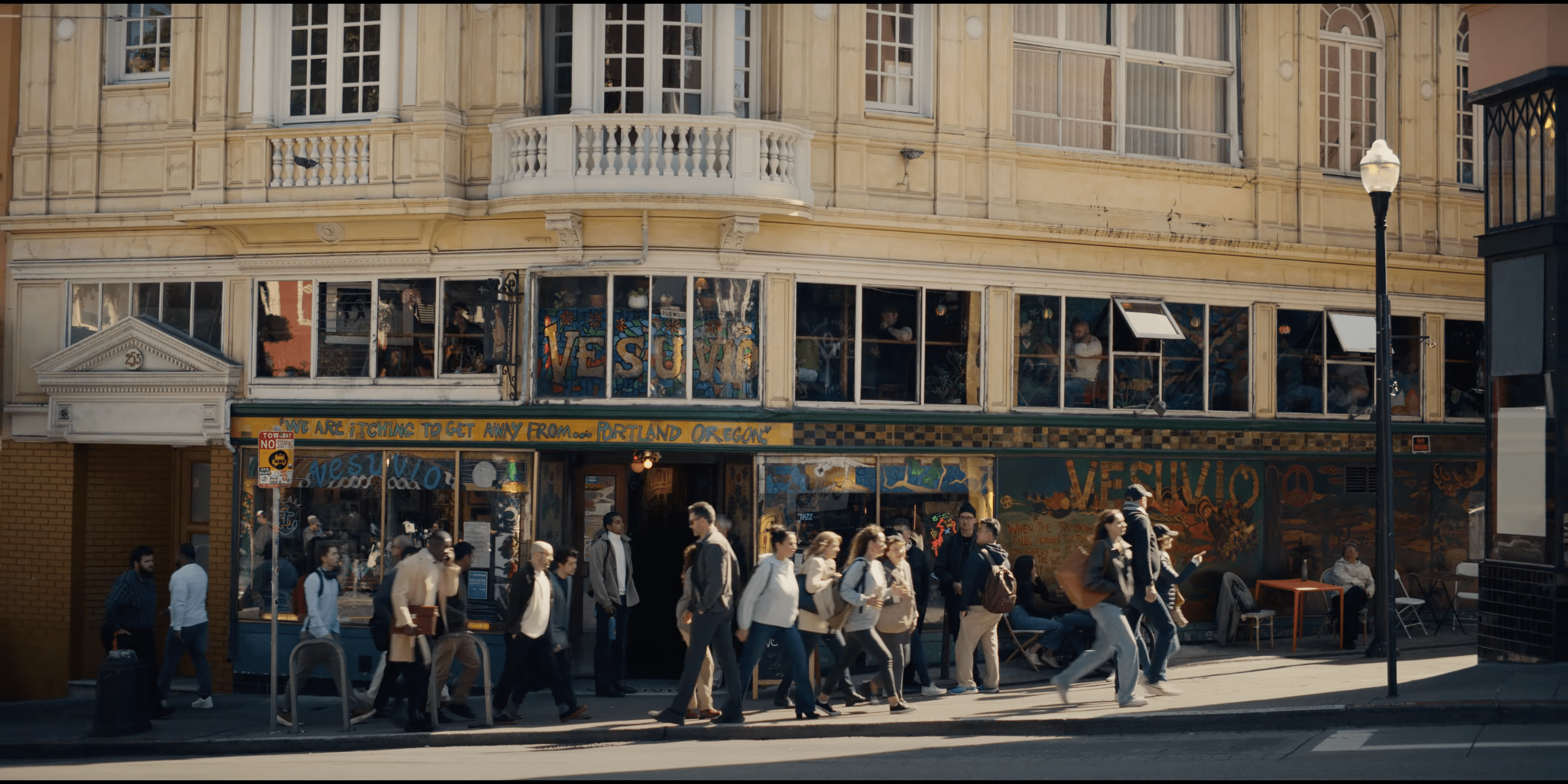 A busy street scene shows a group of people walking past a vintage-looking corner café named &quot;Vesuvio.&quot; The building features large windows and intricate architecture.