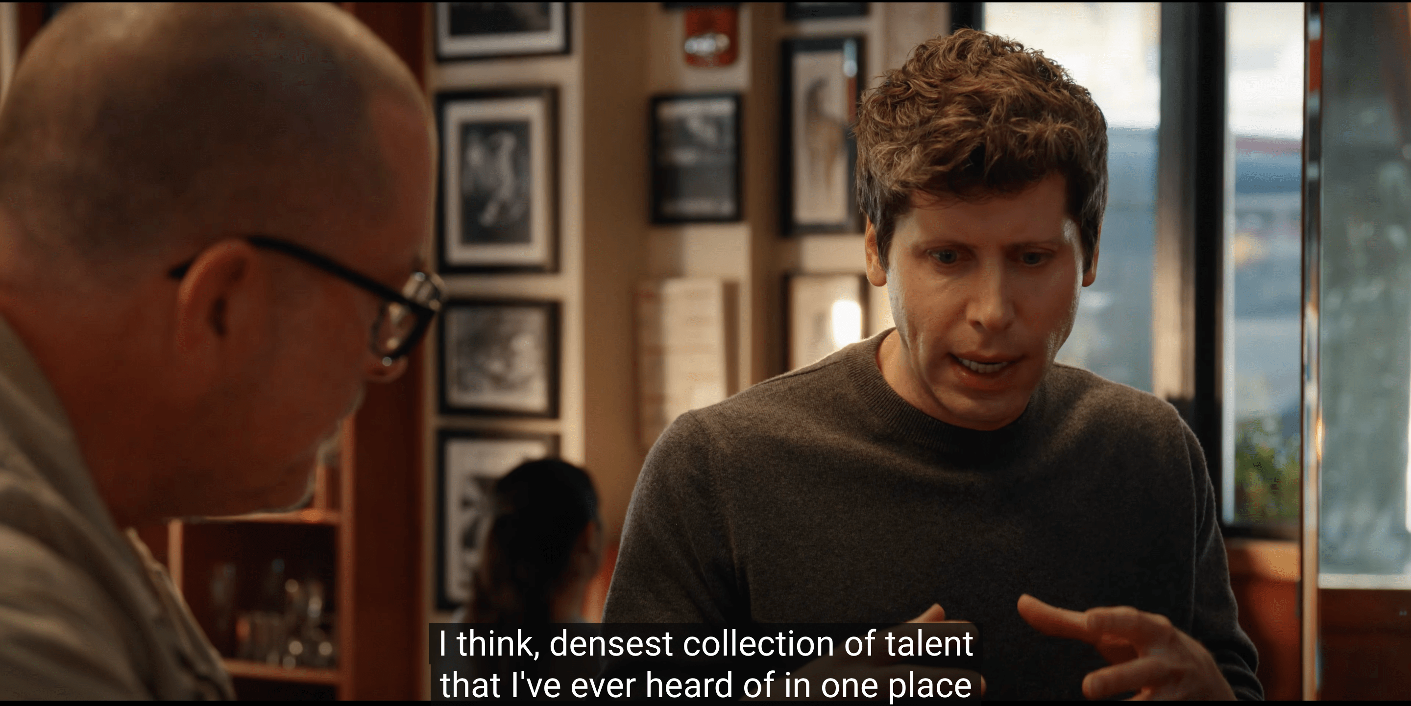 Two men are talking in a room with framed pictures on the walls. One is in profile wearing glasses, and the other faces forward gesturing with his hands. Subtitles read: &quot;I think, densest collection of talent that I've ever heard of in one place.&quot;