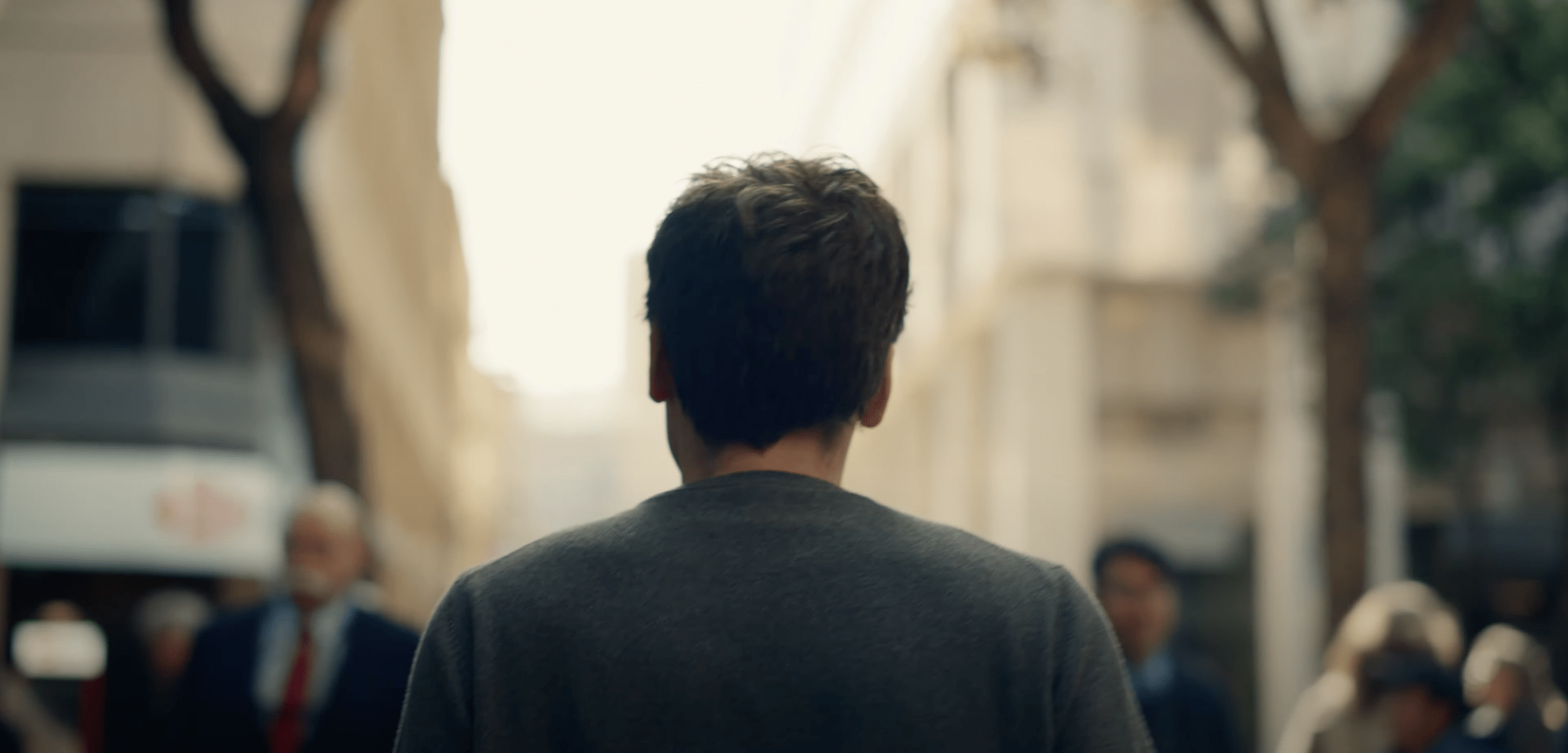 A man with short hair is walking down a blurred city street. He's wearing a gray shirt, and several people are around him, also out of focus.