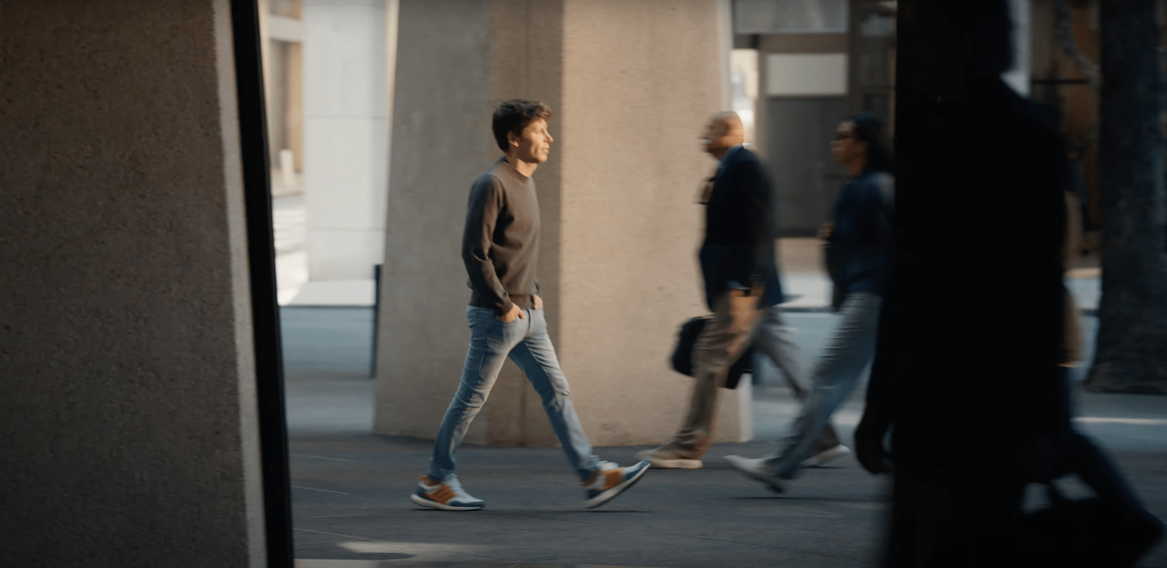 A man walks casually with hands in pockets, wearing a dark sweater, jeans, and sneakers. Others pass by in a blurred, urban setting with tall columns.