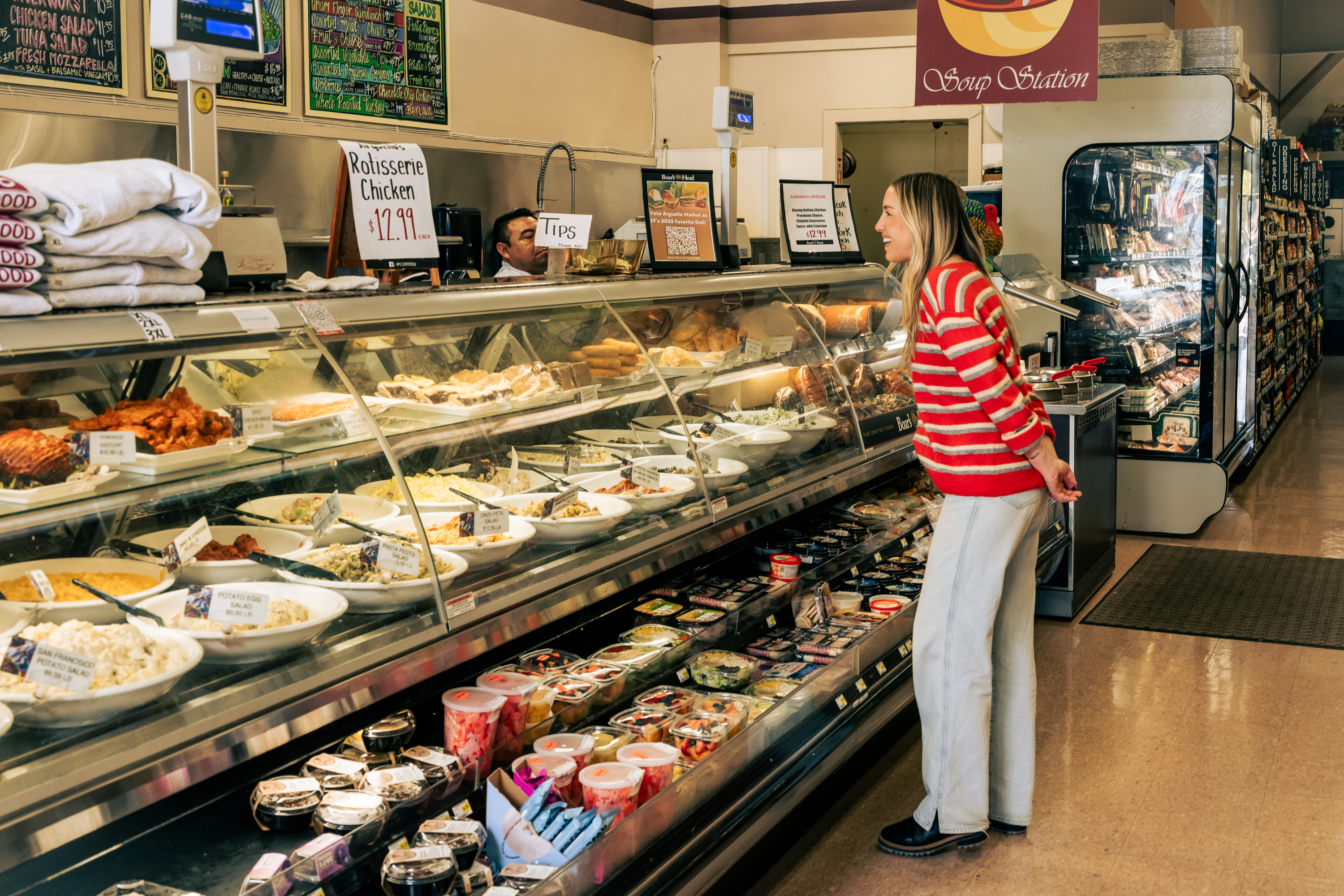 A woman in a red striped sweater examines a deli counter filled with various dishes. The sign above advertises rotisserie chicken for $12.99. The setting is a grocery store.
