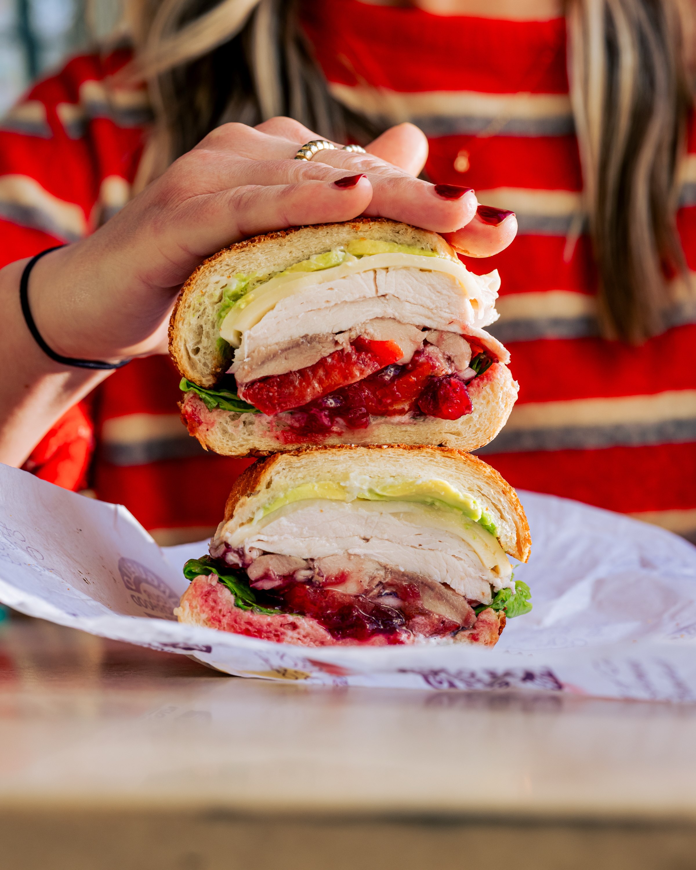 A person holds a halved sandwich with turkey, lettuce, avocado, and red cranberry sauce. The person wears a striped red sweater and has manicured nails.