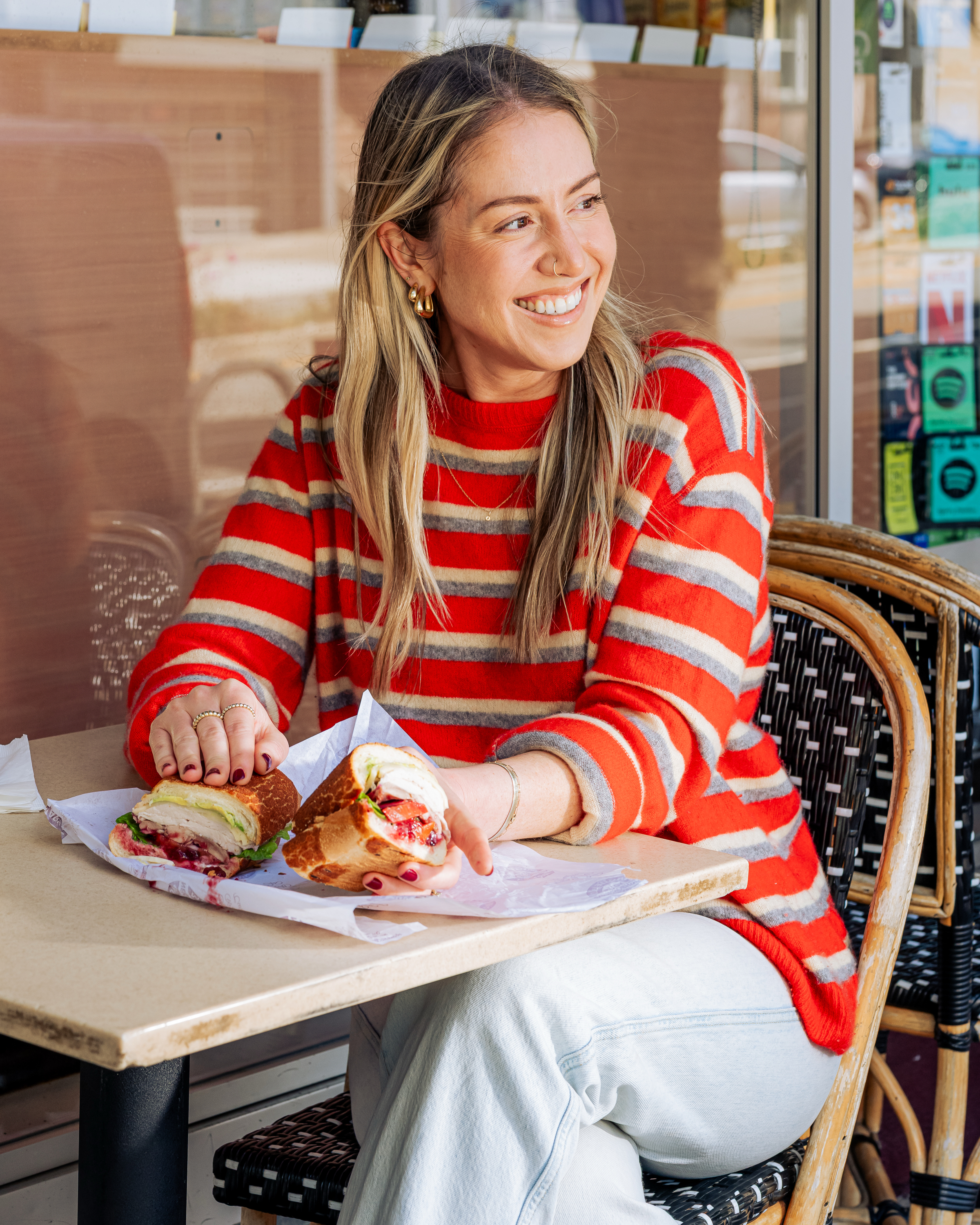 A woman in a red striped sweater smiles while sitting at an outdoor table with a sandwich. She holds one half, looking content and relaxed.