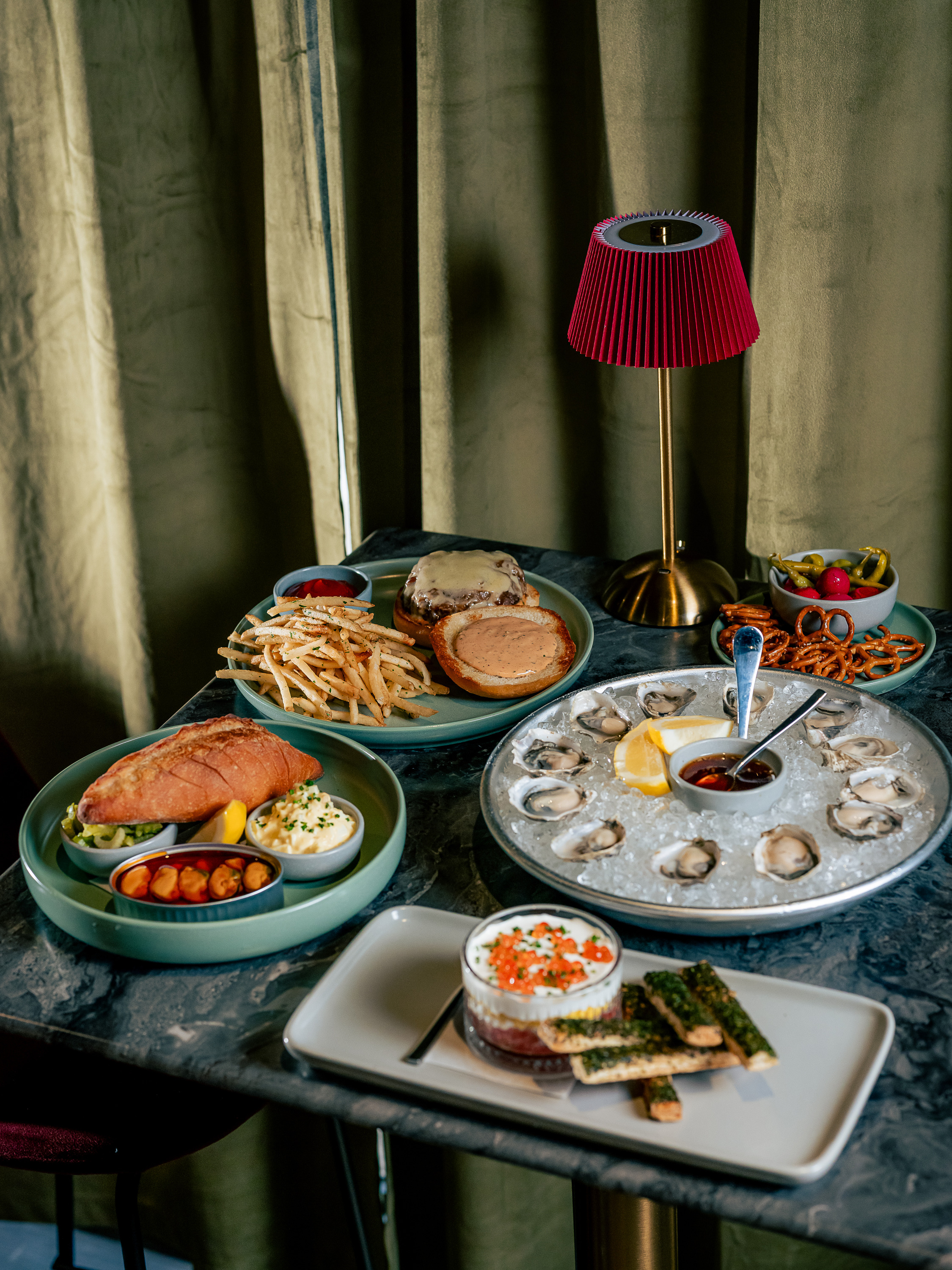 The image shows a table with a variety of foods, including oysters on ice, a sandwich with fries, a burger, a jar with orange toppings, and pickled snacks. A red lamp is in the background.
